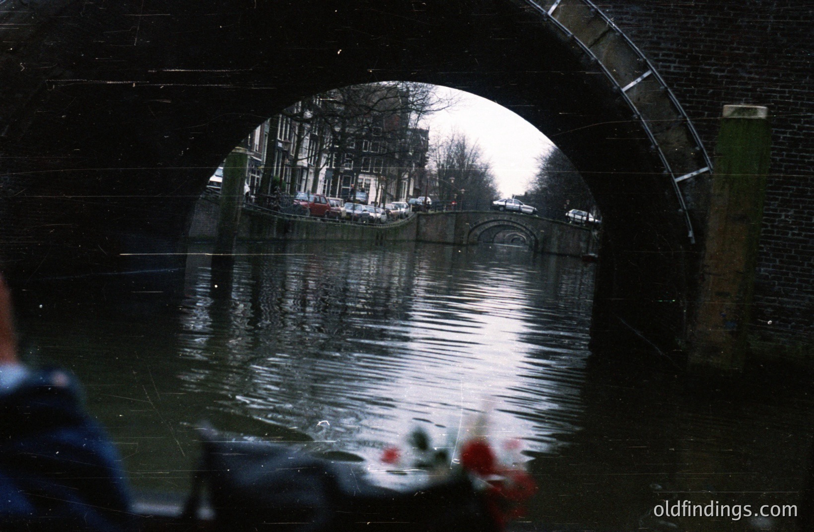 View of a canal scene framed by a brick archway. Traditional Dutch architecture lines the water's edge, reflected in the still surface. Likely Amsterdam, Netherlands. Appears to be a vintage film photograph, exhibiting film grain and slight light leaks. A sense of urban tranquility.