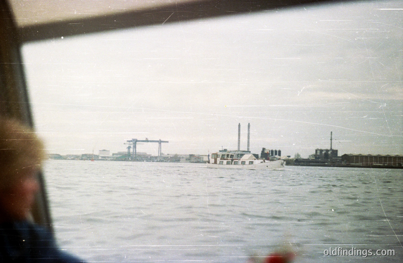 An industrial harbor scene: a boat traverses a body of water, framed by factory structures & cranes along the distant coastline. Likely a port or shipping area, possibly Eastern European. Photograph exhibits wear/aging, indicative of older film stock. Appears to be a tourist or travel image.