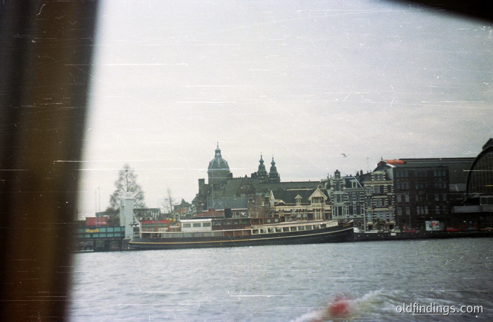 A passenger boat travels along a canal, passing ornate 17th-century Dutch architecture, likely in Amsterdam. The scene is captured through a window, evident by the frame and light leaks. A single bird flies overhead. Potential stock image for travel/heritage themes.