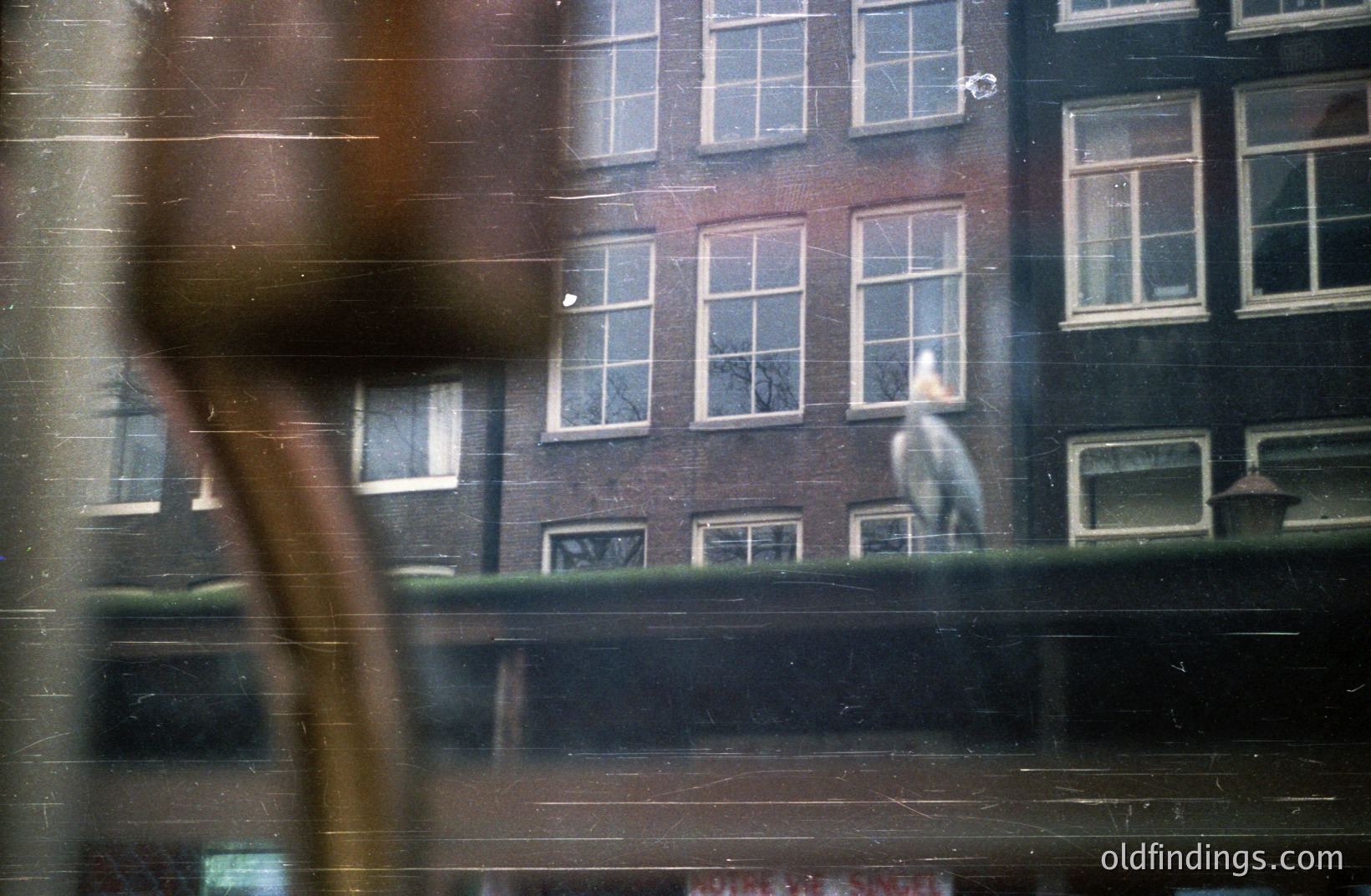 Framed view through windows, a person stands in a brightly lit room with large, multi-paned windows within a red brick building. Appears to be a European urban setting. The image exhibits film grain and light leaks, suggesting a vintage aesthetic, possibly 1970s. A blurred foreground element obscures the lower left.