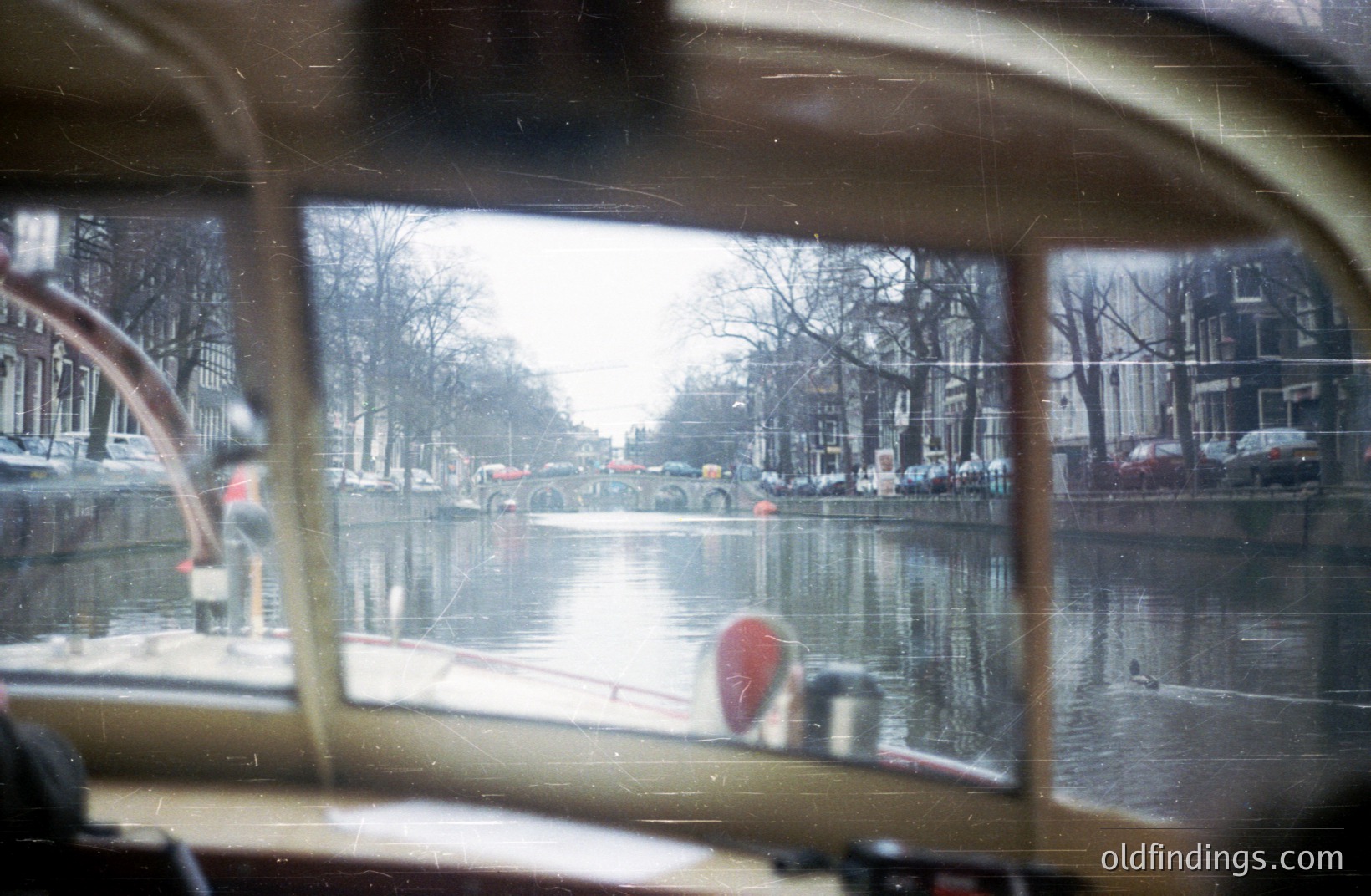 View from within a vehicle, likely a boat, showing a canal scene. Buildings line the waterway, reflecting in the water. Trees are bare, suggesting late autumn or winter. A small red buoy floats in the canal. Likely Amsterdam, Netherlands.