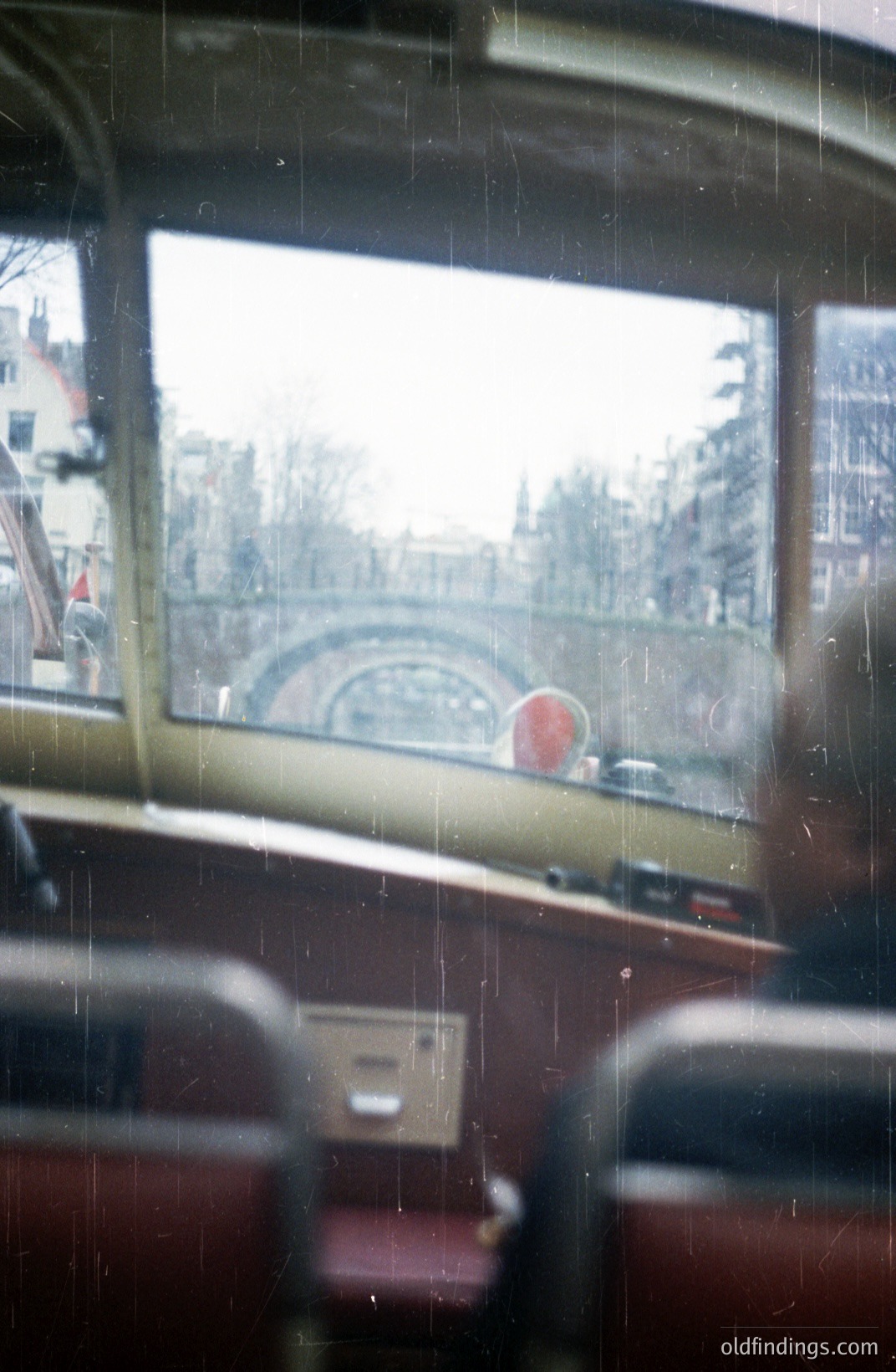 View from a vintage tram, showcasing a canal scene with buildings lining the waterway. Likely a European city, showing a bridge arching over the canal. The interior features red seating and paneling, hinting at mid-20th century transportation. Shows wear and aging.