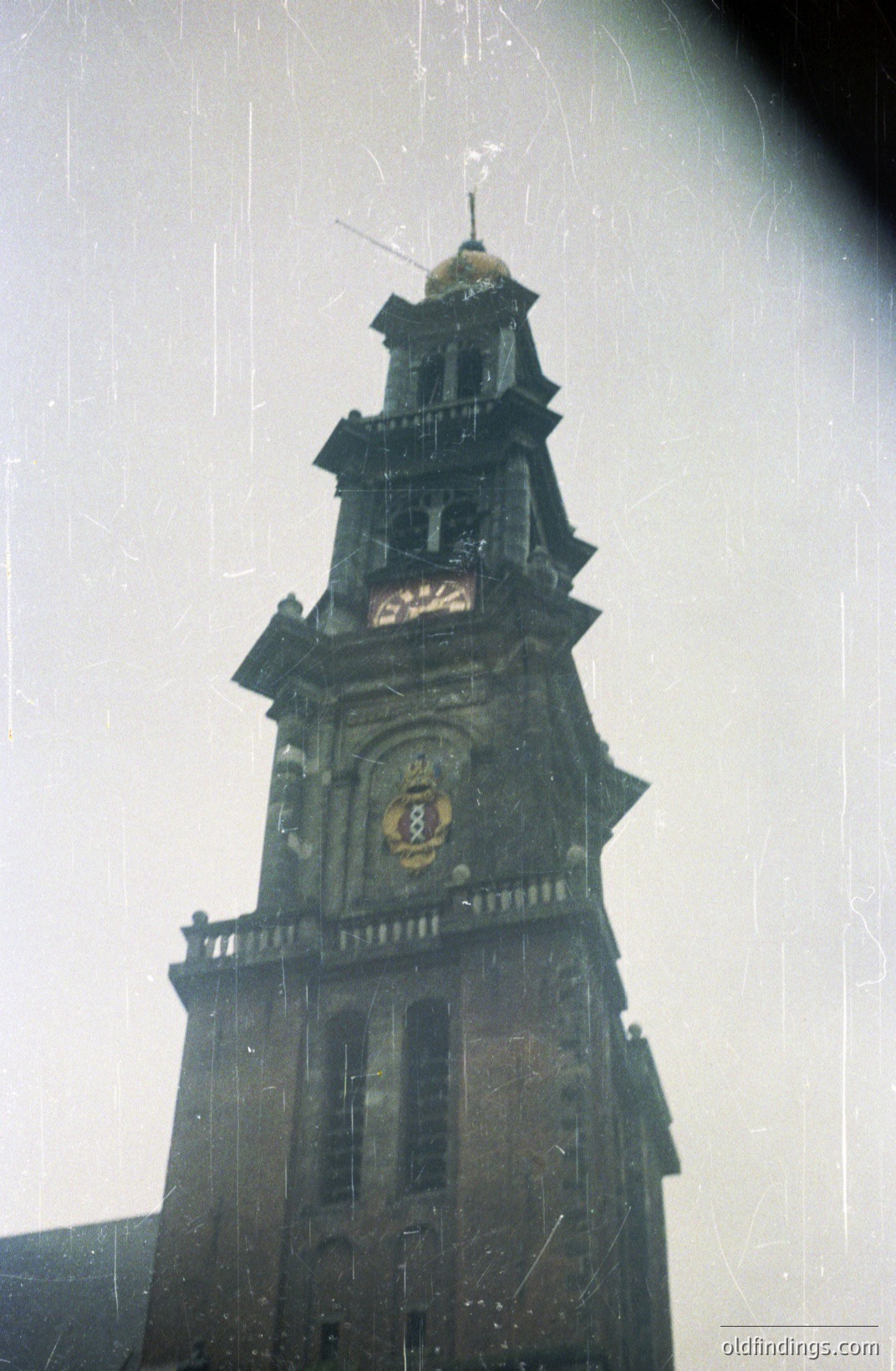 A tall, weathered stone church tower dominates the frame, featuring a clock face and ornate detailing. Visible signs of aging and environmental exposure, including discoloration and surface wear. Likely European architecture, potentially 18th or 19th century. The image exhibits film grain and light leaks.