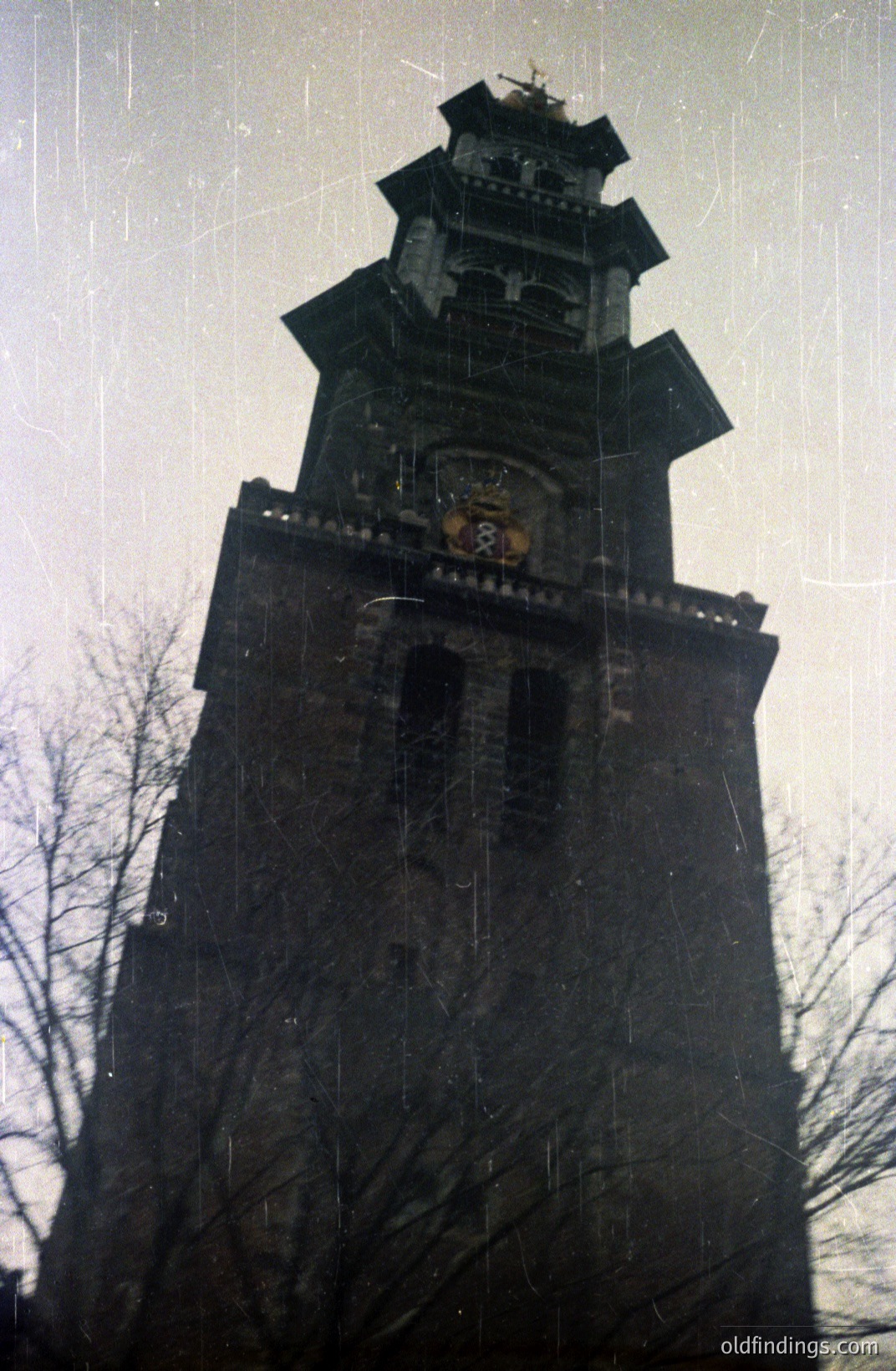 A tall, multi-tiered brick tower with decorative stonework and a pointed roof dominates this photo. A crest or emblem is visible above the central section. The aged film stock displays considerable grain and scratches. Likely a European structure, possibly 20th century.