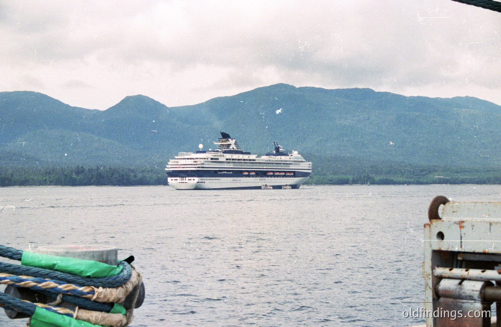 A large cruise ship cuts through a calm body of water, backed by a backdrop of forested mountains under an overcast sky. Ropes and metal railings frame the foreground view. Likely a coastal scene, possibly Scandinavia or similar northern latitude. Appears to be a snapshot, potentially from the 1990s or 2000s. Useful for travel or nautical design references.