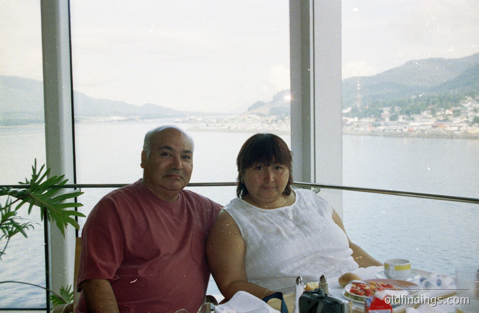 A couple sits at a table, facing the camera, with a scenic coastal view visible through large windows. The view shows a town nestled at the base of a mountain range. Likely a vacation photo, the style suggests the 1980s or 1990s. Simple composition, potentially useful as a nostalgic travel reference.