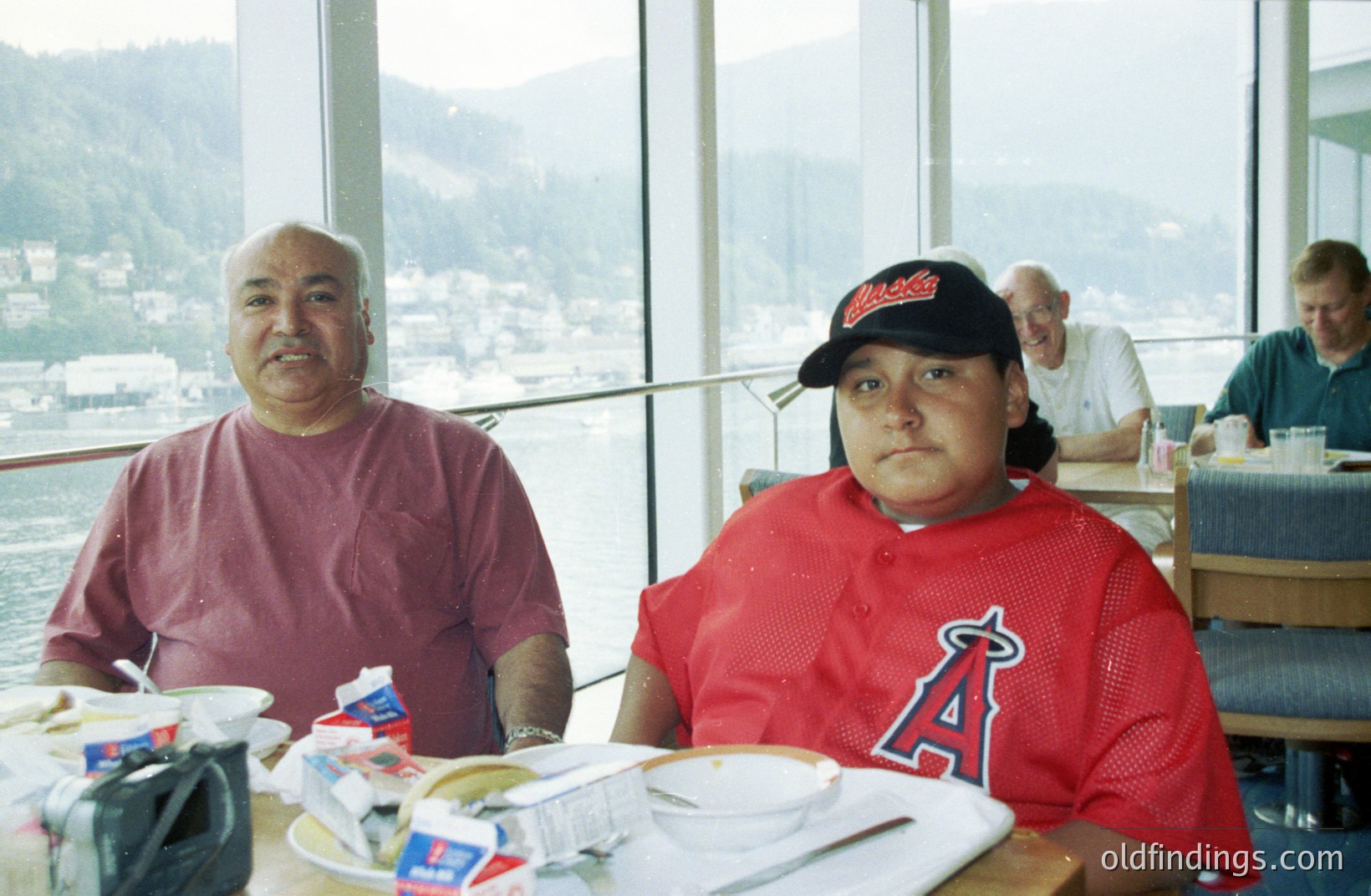 Two men seated at a table by large windows overlooking a coastal landscape with mountains in the distance. The man on the left wears a red sweater, the other an Anaheim Angels baseball jersey & hat. Tray with food visible on table; potentially a cruise ship dining room. Likely 1990s or 2000s.