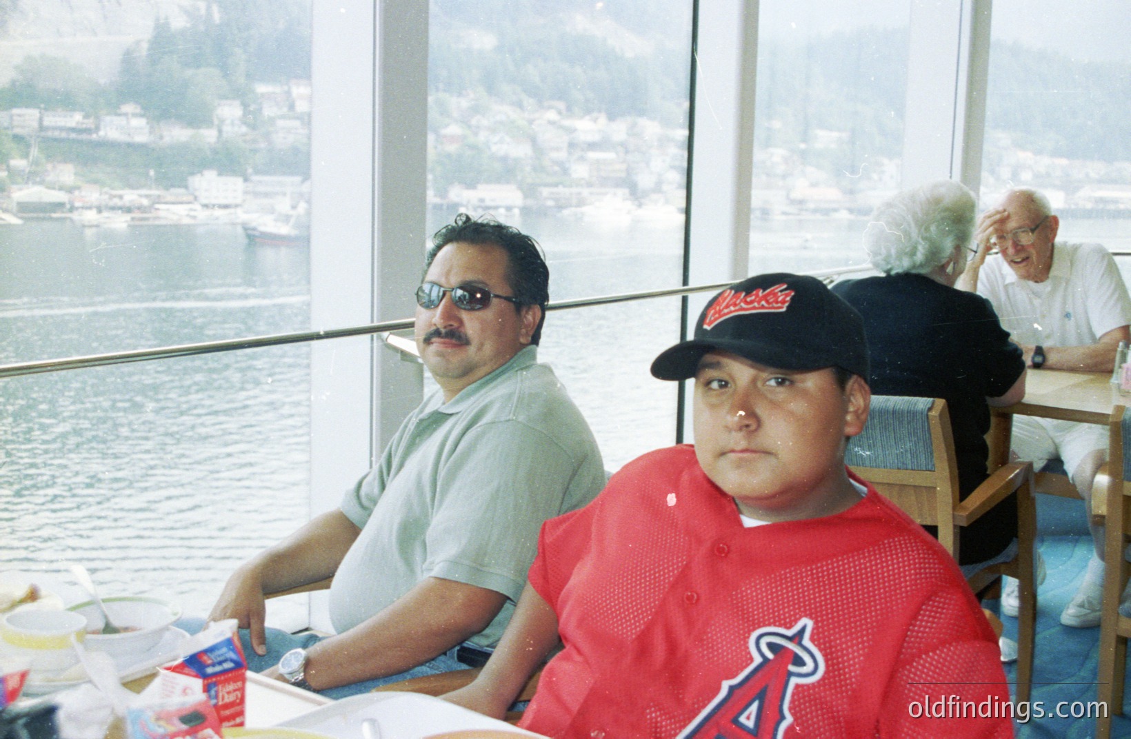 Two men seated at a restaurant with large windows overlooking a harbor and mountainous coastline. One wears sunglasses and a light green shirt, while the other has a baseball cap and red Anaheim Angels jersey. A partially visible older man and woman are seated nearby. Likely 1990s, coastal location.