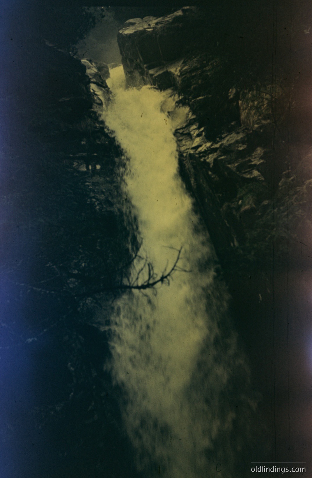 Striking sepia-toned image captures a cascading waterfall amidst dark, rocky terrain. A lone, bare branch juts diagonally across the falls. Likely a vintage photograph, the image exhibits signs of age and handling – surface scratches and discoloration. Evokes a sense of isolation and timelessness.