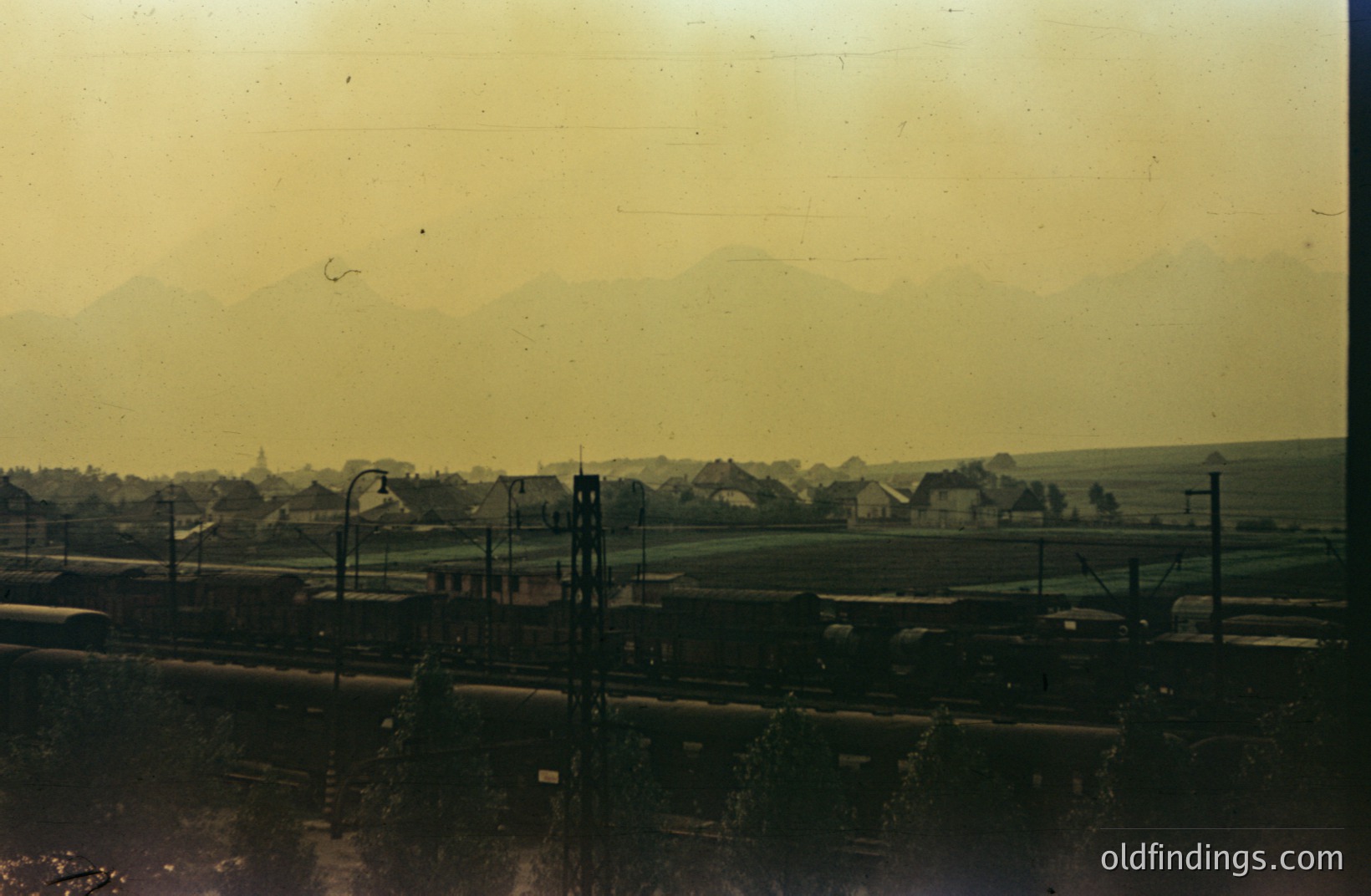 A color photograph depicts a landscape with a railway line in the foreground and distant mountains in the background. Several train cars are visible on the track. A town or village is set against the mountains, with a church spire visible. Heavy atmospheric haze impacts color and clarity. Appears to be 1960s-1970s.