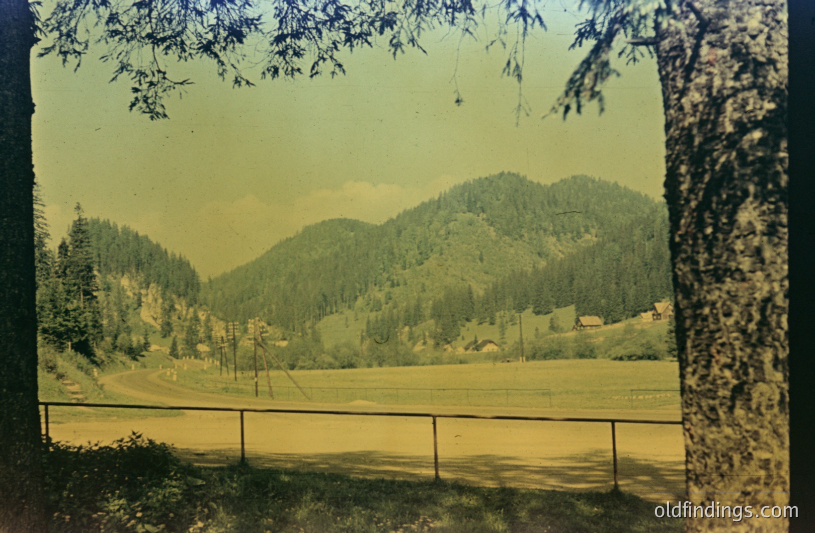 A wide, expansive view across a grassy field toward forested mountains. A section of dark wooden fencing runs along the foreground. The scene is framed by tree branches in the upper corners. The photograph's color palette suggests a vintage film print, possibly 1950s-1970s. Likely a recreational area or park setting.
