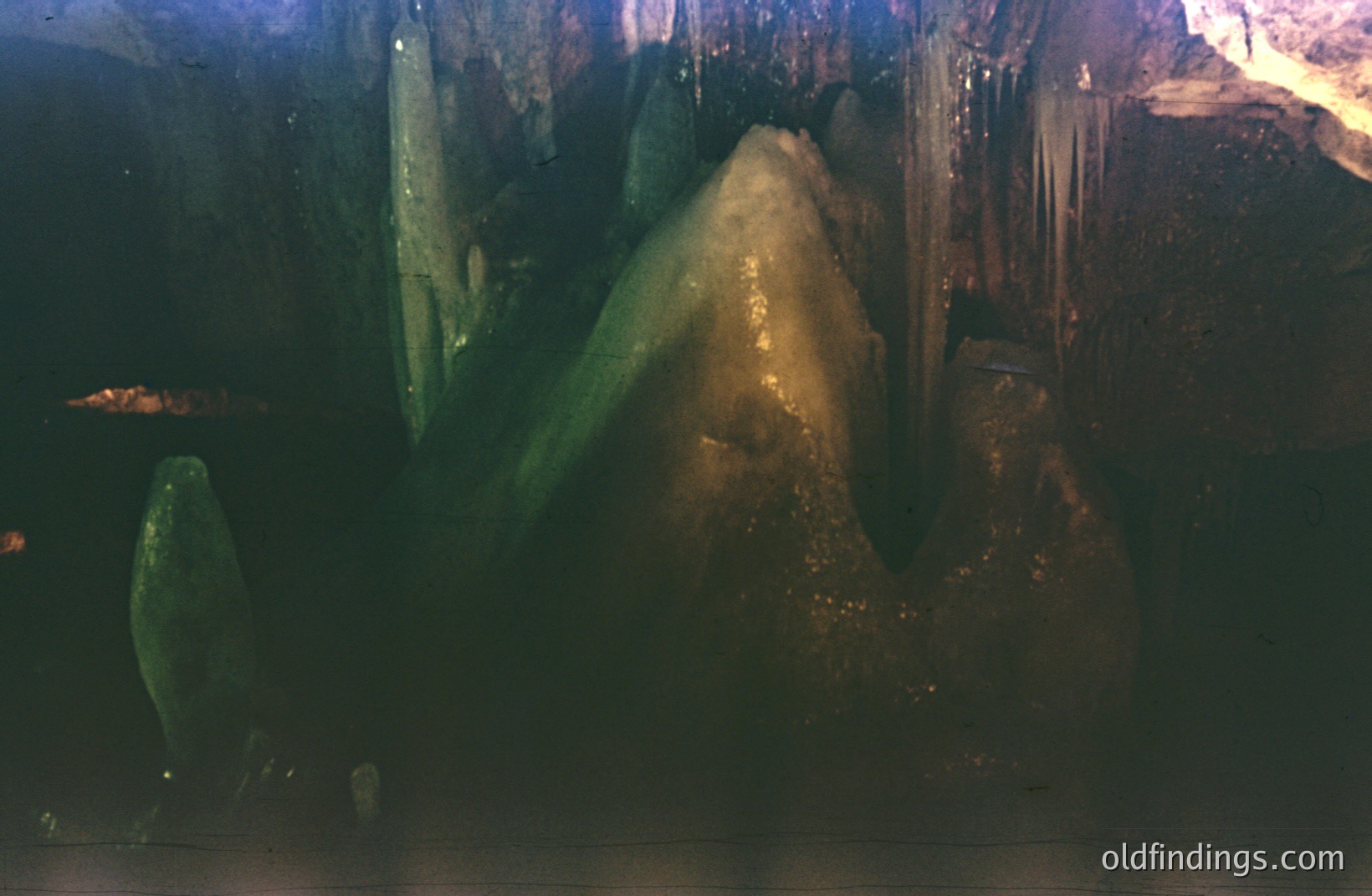 A view within a grotto or cavern, showcasing textured rock formations illuminated by dramatic, directional lighting. Visible stalactites and stalagmites suggest a limestone cave environment. The color palette leans towards deep greens and browns, hinting at an aged, natural setting. Likely 1970s era, possibly intended for travel or educational stock imagery.
