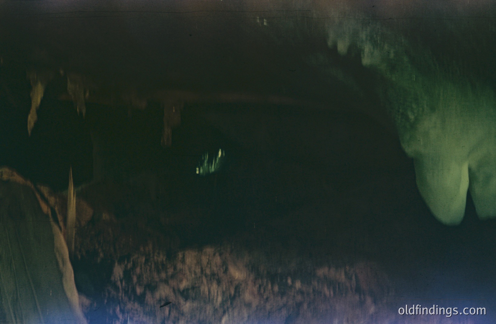 A wide, dark cave interior. Limestone formations, likely stalactites and stalagmites, define the cavern walls. A narrow slit reveals a glimpse of bright sky/water, creating a dramatic contrast. The texture is rough and damp, highlighting the natural geological processes.
