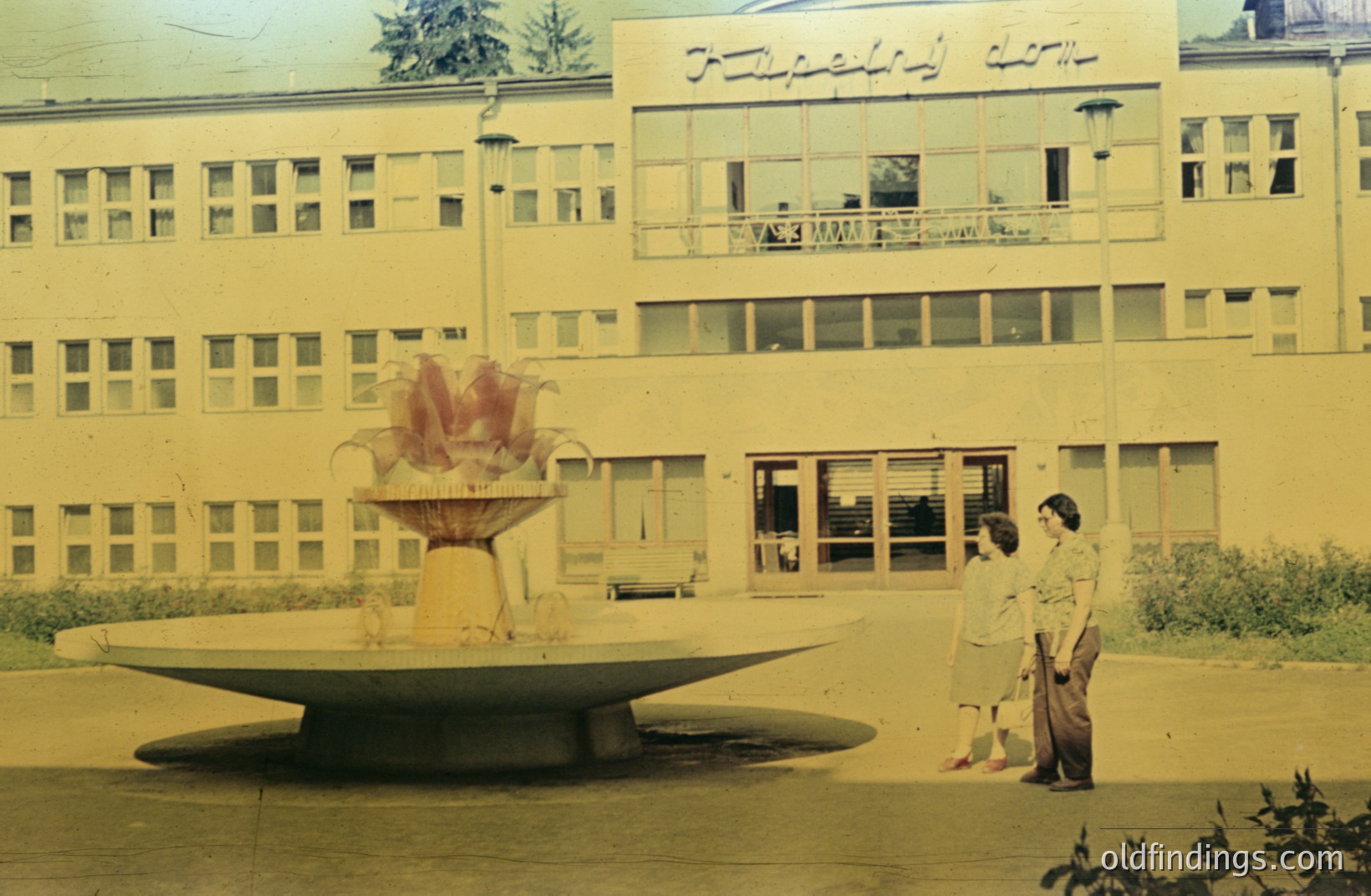 A wide, tiered fountain dominates the foreground of a vintage photograph. A modern, multi-story building with a grid of windows forms the backdrop. Two figures stand on a walkway near the fountain. Appears to be a public space, possibly a university or civic building. Architectural style suggests 1960s or 1970s.