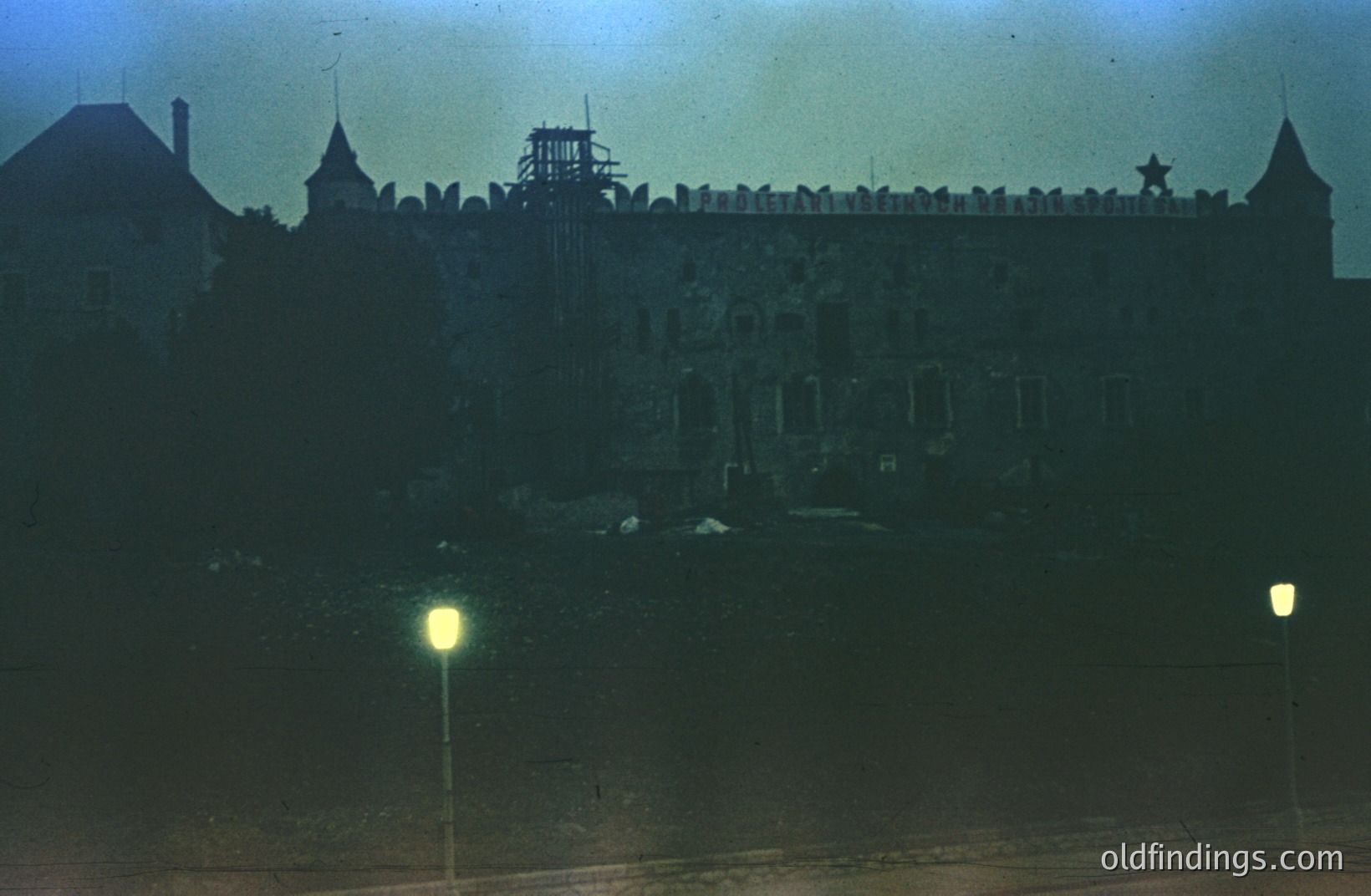 Striking low-angle view of a monumental, austere building featuring a central tower topped with a star. Likely a government or institutional structure. Visible signage in Cyrillic script. The austere architecture and style suggest a mid-20th century (1960s-1970s) Eastern European aesthetic. Two lampposts illuminate the foreground.
