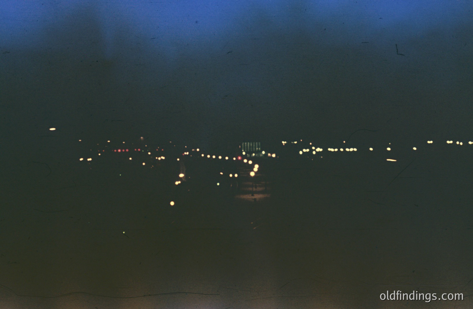 Long-exposure view of a string of lights along a shoreline, possibly a pier or walkway, with what appear to be buildings illuminated in the distance. The effect creates streaks of light. A dark, overcast sky dominates the scene. Likely taken during the 1960s or 1970s, judging by film characteristics.
