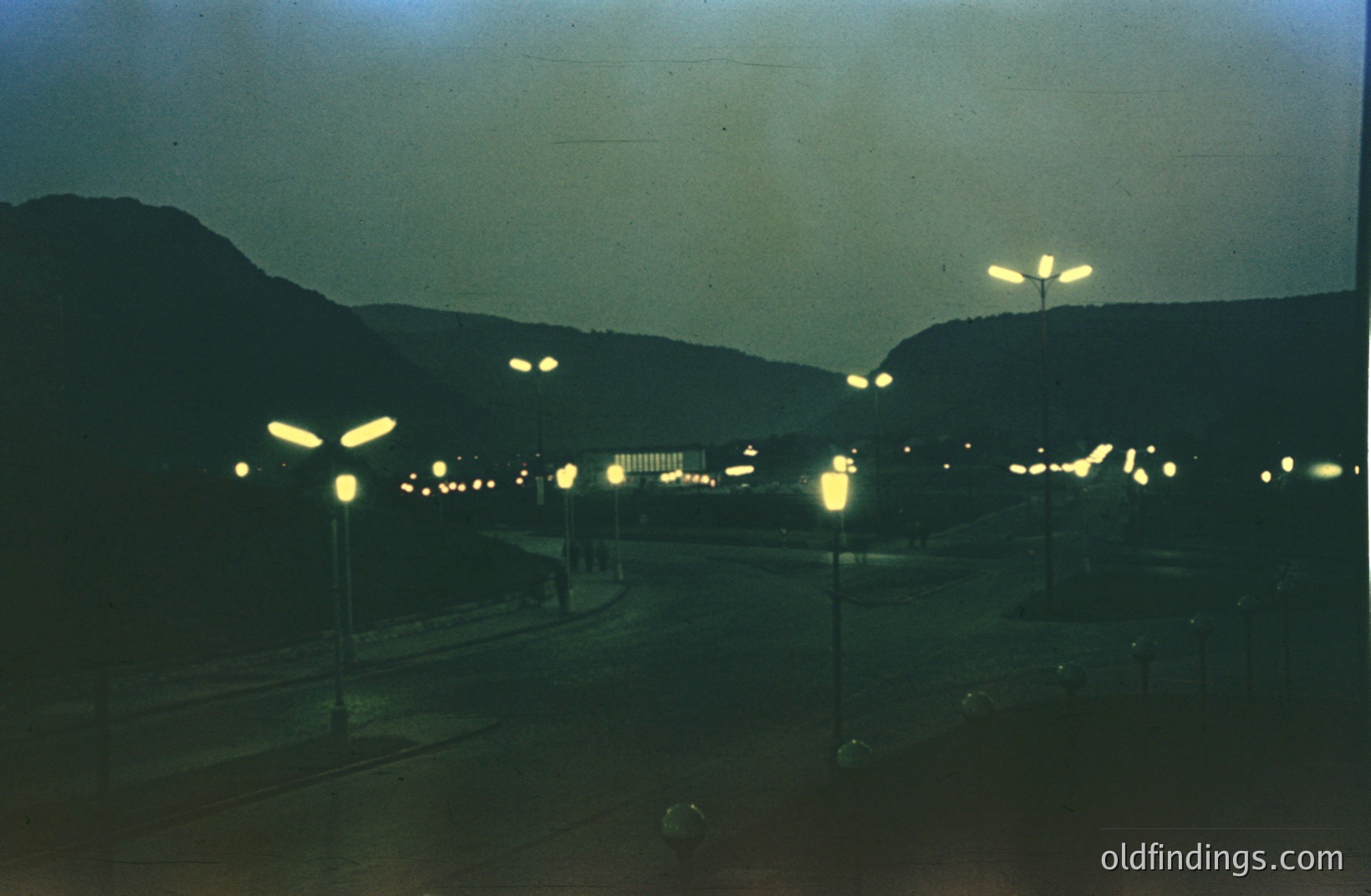Night view of a town nestled within a mountainous landscape. Prominent, stylized lampposts illuminate a wide plaza or courtyard. The architecture displays mid-century modern influences. Likely Eastern European, possibly 1960s-1970s. A potential stock photo reference for urban planning or design.