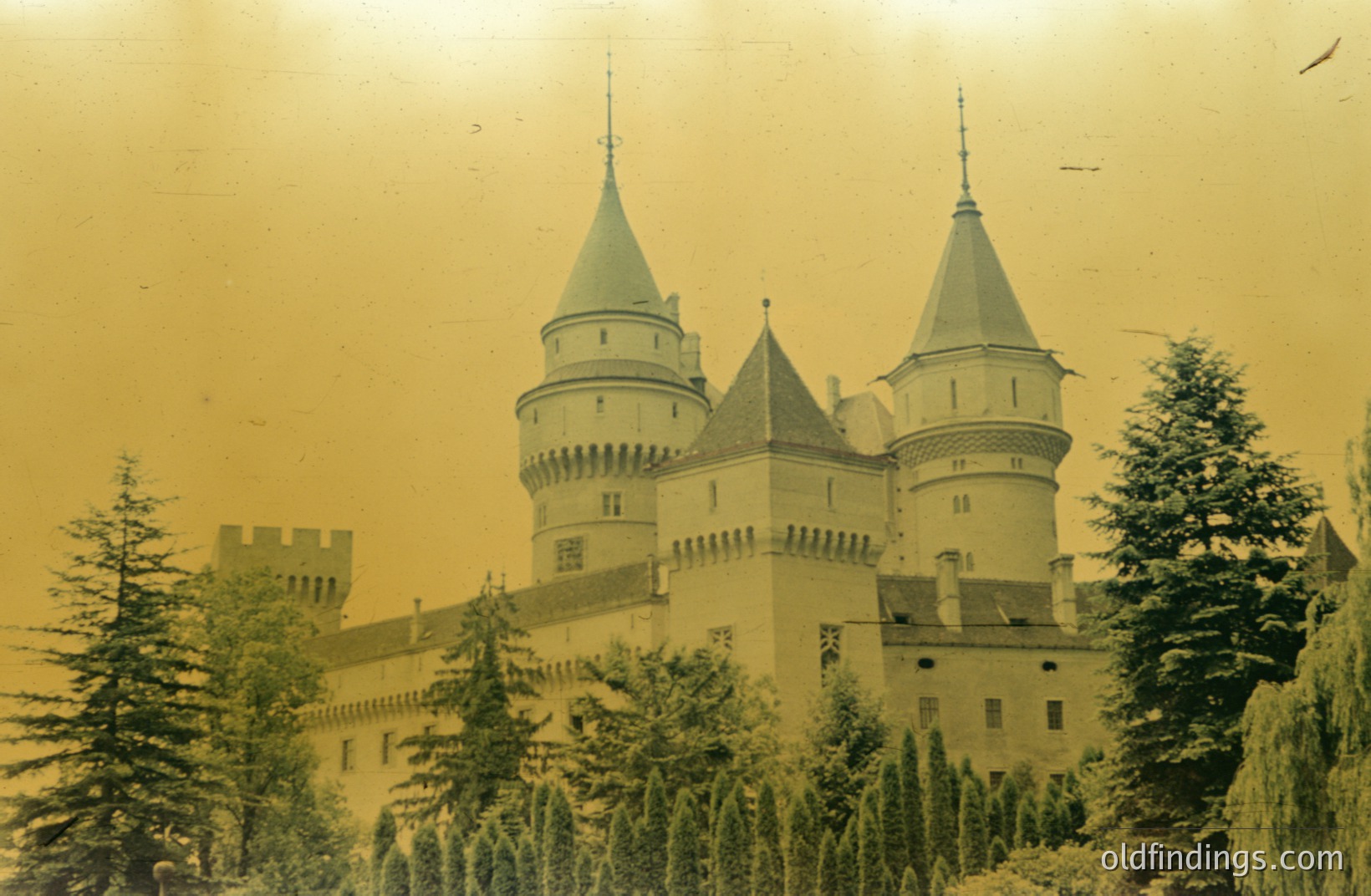 An imposing castle, likely Bavarian or Austrian, features prominent corner turrets and pointed spires. Dense evergreen trees frame the structure, creating a picturesque alpine scene. Sepia toning suggests a vintage photograph, potentially 1930s-1950s. Represents architectural grandeur.