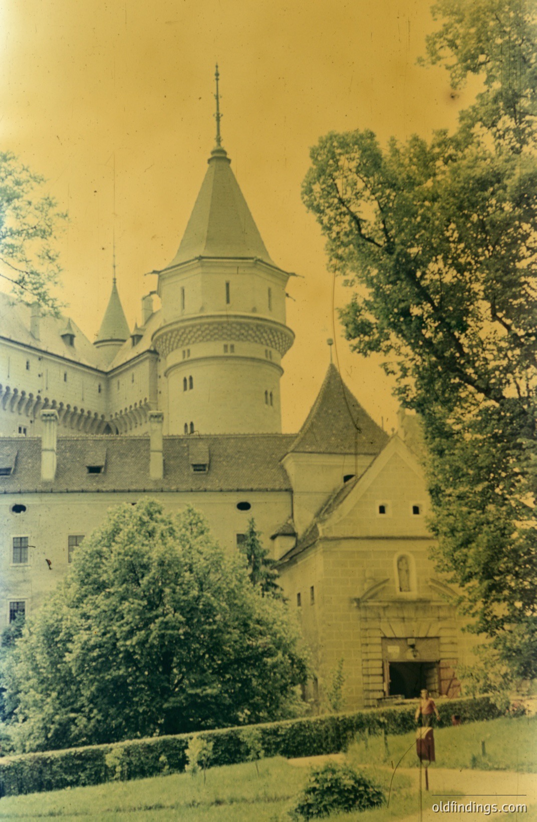 Stunning view of a castle complex, likely European, framed by lush greenery. Stone architecture features turrets, a central cone-shaped tower, and a walled entrance. Appears to be an early color photograph, perhaps 1950s-1970s. A single figure stands near the gate. Potential for stock imagery & heritage research.