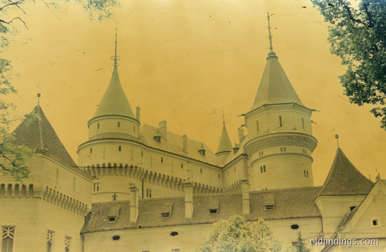 A vintage, sepia-toned photograph depicts a stately castle with prominent conical towers and crenellated battlements. Architectural style suggests late medieval or early Renaissance influence. Likely European, possibly Central or Eastern Europe. The aged print exhibits signs of wear and fading.