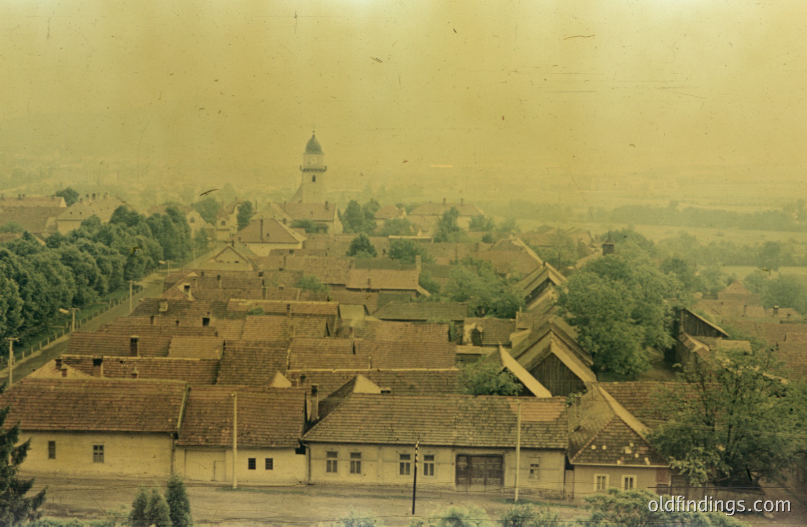 A high-angle view showcases a European village with tightly packed, red-tiled roofs. A church steeple rises above the buildings, framed by lush greenery. The image exhibits a vintage, faded color palette, likely dating from the mid-20th century. A classic architectural record.