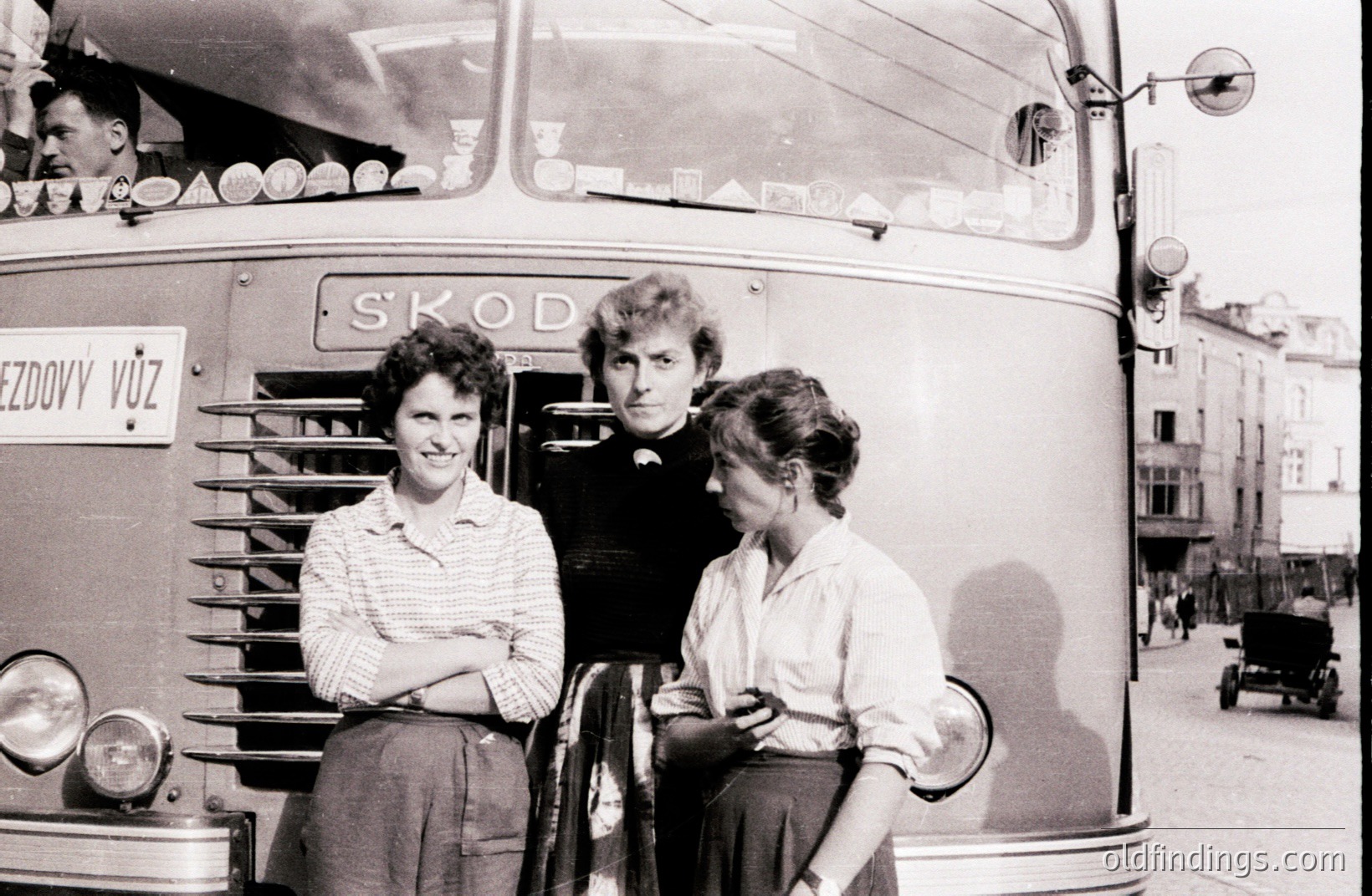 Three people pose by a Škoda bus, likely in Czechoslovakia (now Czech Republic). The bus displays "PŘEZDOVY VŮZ" signage. Two women in modest dresses and one man with dark hair, captured in a candid, snapshot style. Possibly 1960s street scene with period architecture and clothing. #Škoda