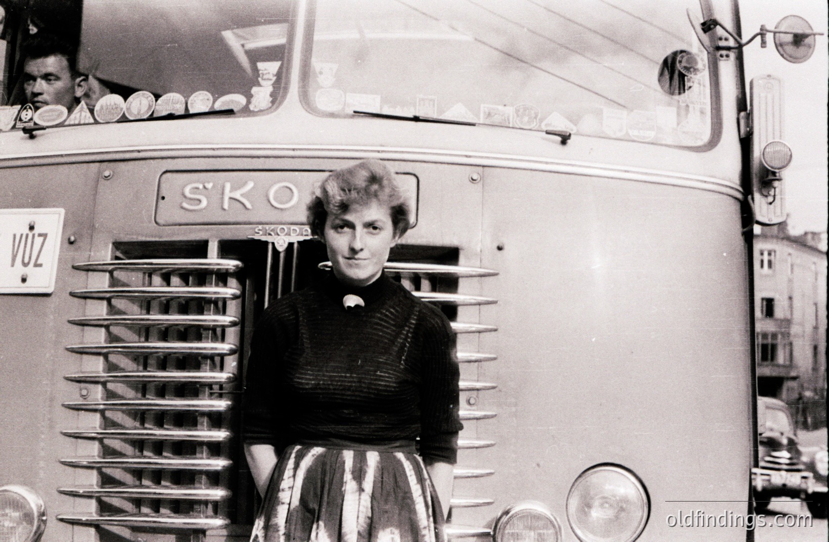 A young woman stands proudly beside a Škoda bus, likely in Czechoslovakia. She wears a dark, textured sweater and a full, patterned skirt, indicative of the 1950s-60s fashion. The bus features distinctive chrome detailing & a "VÚZ" plate. A driver is visible within. #Škoda