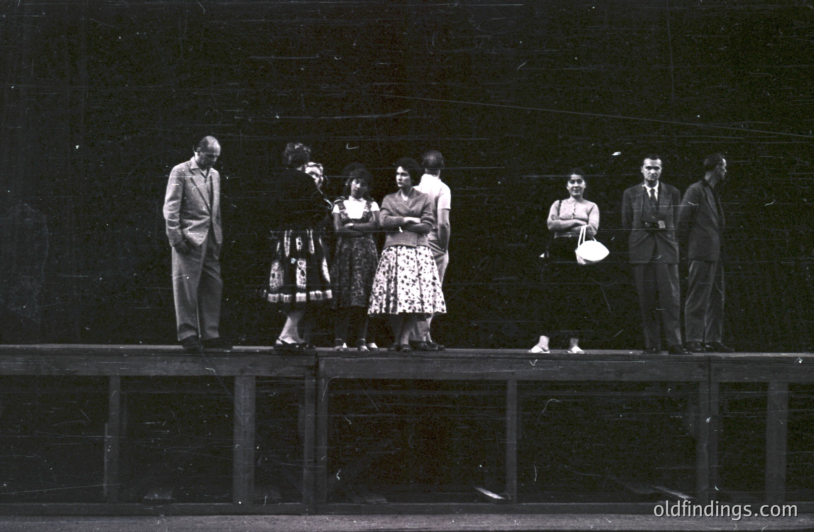 Group of seven individuals, six adults and one child, stand on a raised platform or stage, facing the camera. Their attire suggests the 1960s or 1970s – tailored suits, dresses, and modest footwear. Likely a theatrical or performance context, stark lighting emphasizes the figures against a dark backdrop.