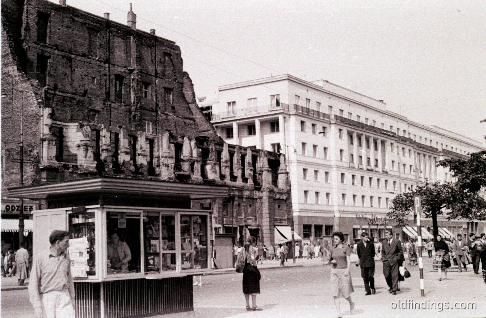 Wartime destruction contrasts with the intact, modernist apartment building façade. A damaged structure exposes brickwork and roof framing. Pedestrians stroll past a kiosk & along the street. Likely Sofia, Bulgaria, post-WWII, 1940s-1950s. Historical record of urban recovery.