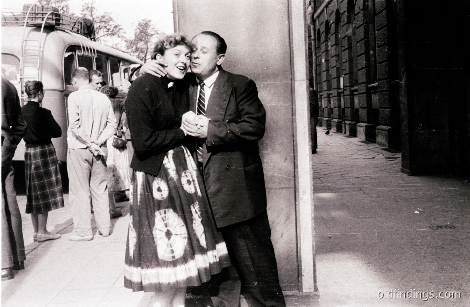 A smiling couple embraces near a building exterior, likely at a transit station. The woman wears a patterned full-skirted dress, while the man is in a dark suit and tie. Background shows people and a bus. Likely 1950s. A snapshot capturing a joyful moment.
