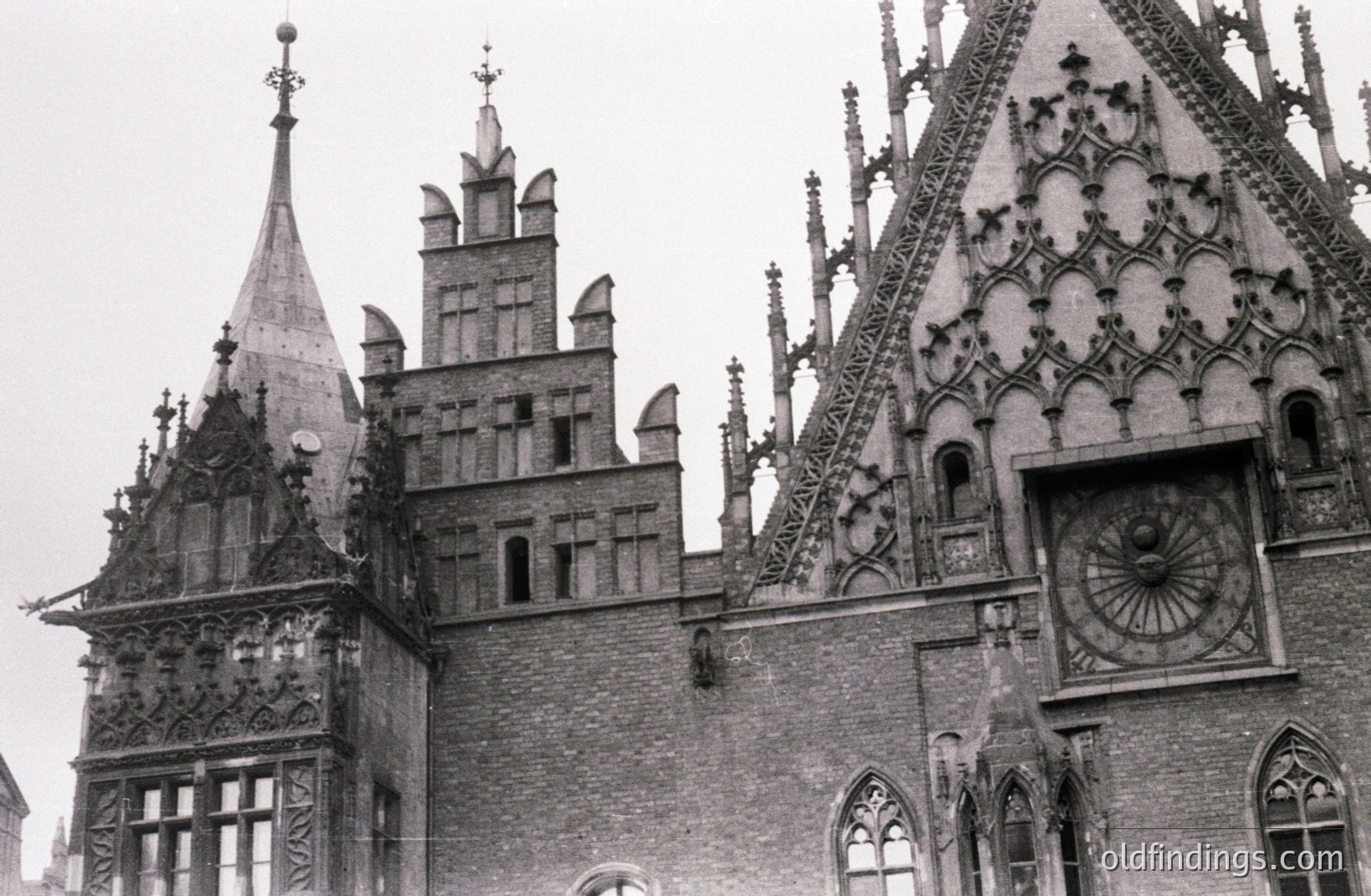 A black and white photograph showcases intricate Gothic Revival architecture. A central spire rises above a complex brick facade adorned with decorative tracery and pointed arch windows, including a large sundial. Likely European, dating from the late 19th or early 20th century. Demonstrates craftsmanship and design motifs.