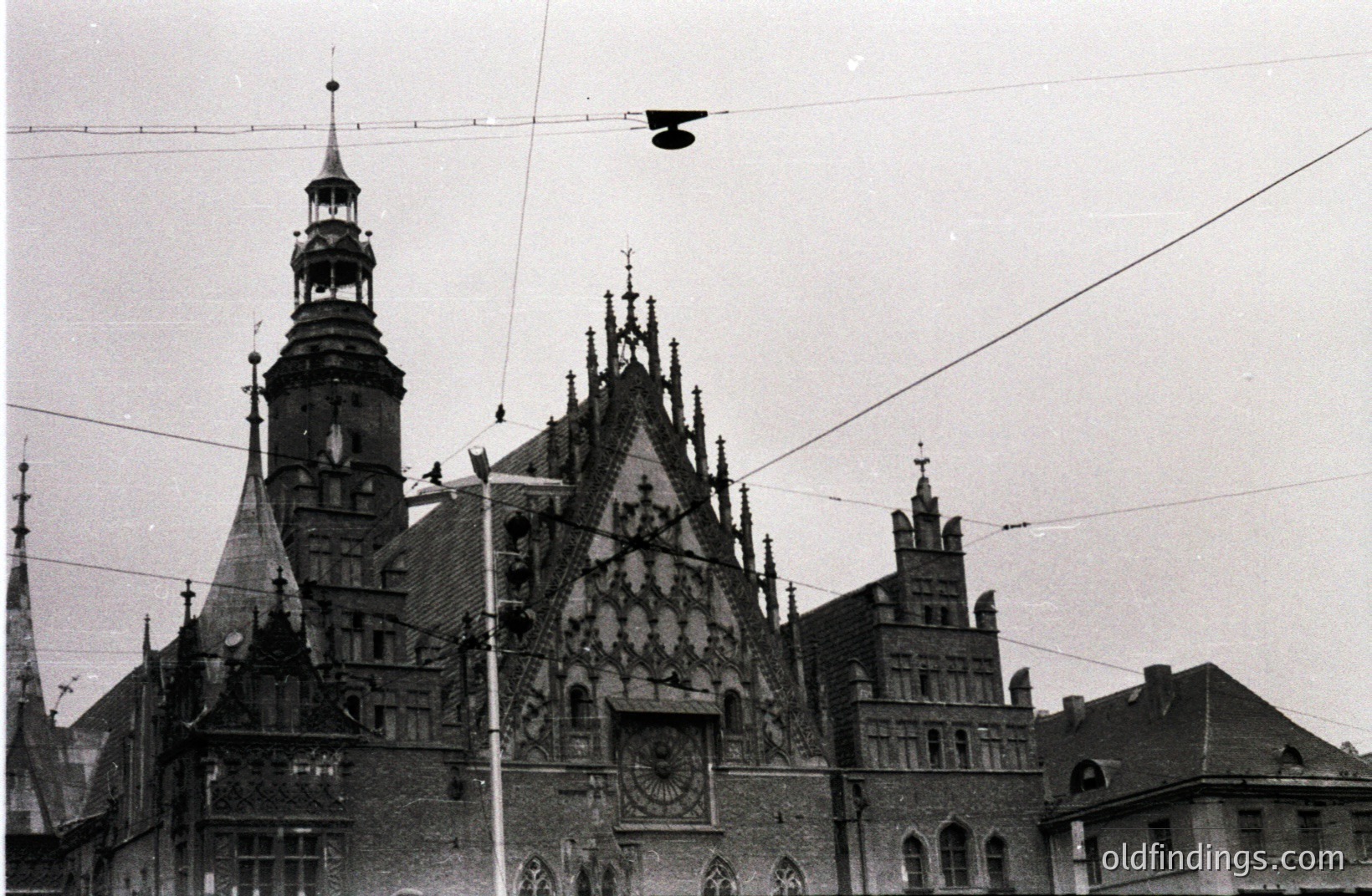 Ornate, historic city hall with a towering clock and detailed, peaked roofline. The building displays Gothic Revival architecture, likely from the 19th century. Visible signage suggests a European location, possibly Poland. Power lines cross the sky. Excellent reference for architectural studies.