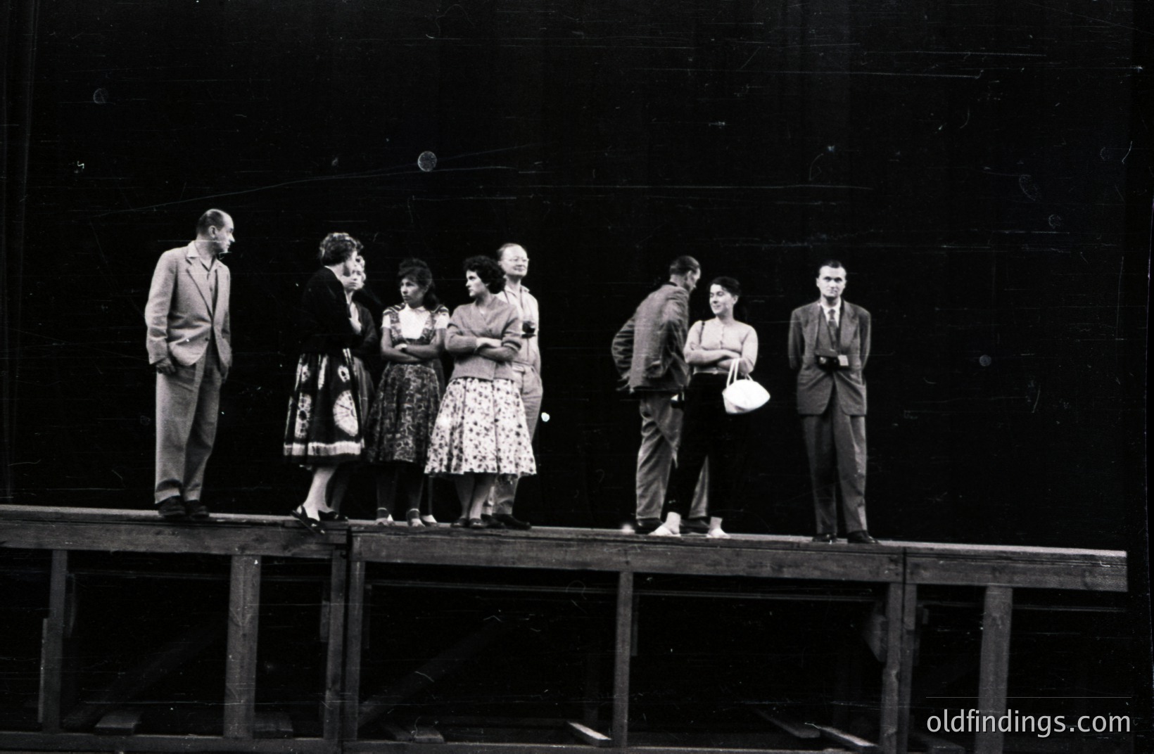 Six individuals stand on a wooden platform against a dark, possibly stage backdrop. Men in suits and a woman in a dress appear alongside younger individuals in casual attire. Likely a theatrical production still or behind-the-scenes shot. Appears to be from the 1950s-60s. A sense of formality contrasted with relaxed poses.