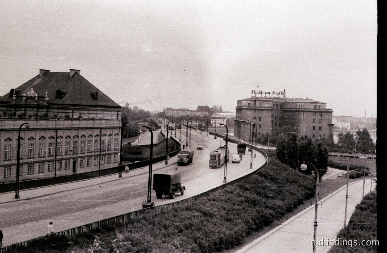 An elevated, wide avenue curves into the distance, featuring early automobile and truck traffic. A monumental, classically-styled building dominates the left foreground; another, taller structure stands on the right. Likely post-war reconstruction, possibly Eastern Europe, 1950s-1970s. Architectural details suggest socialist realism.
