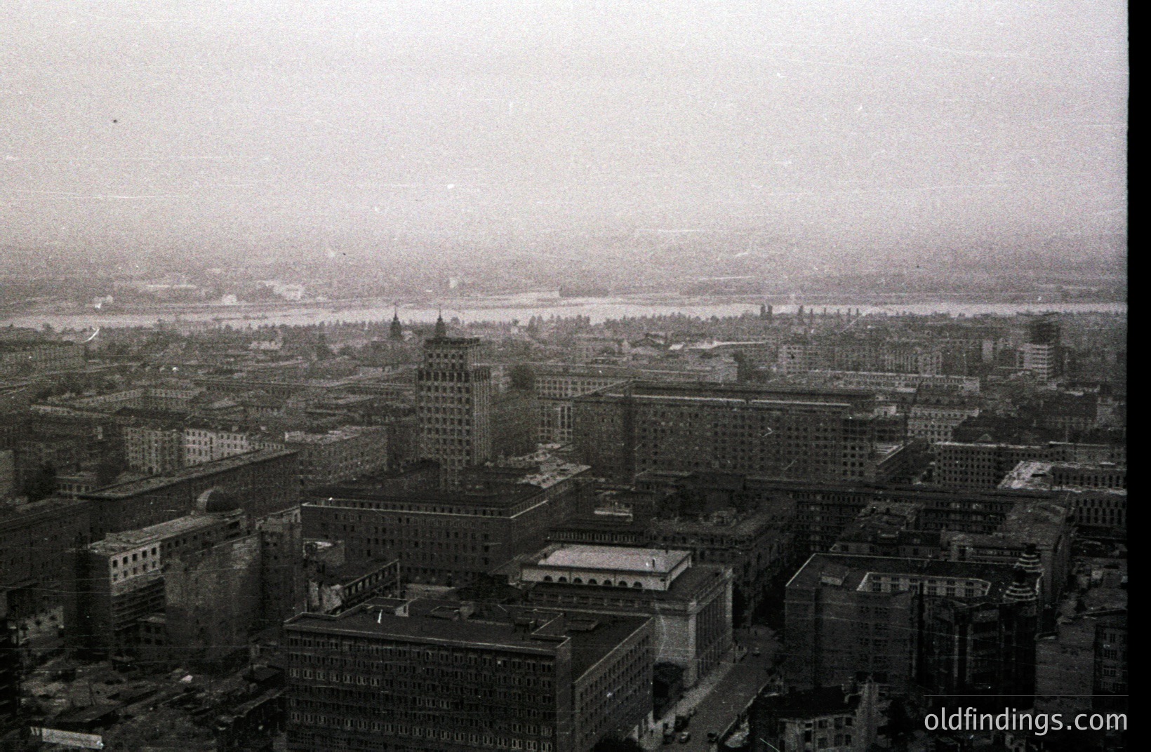 A high-angle, monochrome cityscape shows dense urban architecture along a wide river. Buildings display early 20th-century design, featuring ornate facades and varied heights. The river recedes into the hazy distance. Likely taken in the 1920s-1940s, possibly New York City. Historic urban planning and design reference.