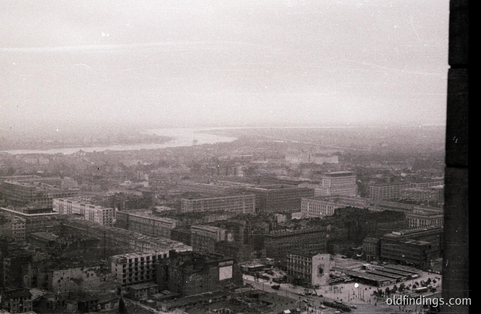 High-angle view of a cityscape featuring dense, mid-century architecture. A wide river dominates the background, partially obscured by atmospheric haze. Buildings exhibit a functionalist style, typical of post-war reconstruction. Likely European, possibly 1950s-1970s. Potential stock photography value for architectural studies.