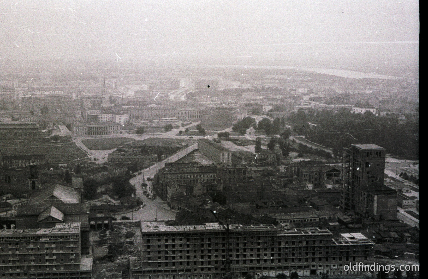 High-angle cityscape view. Extensive urban sprawl with a prominent central plaza & institutional buildings. Visible river in the distance under a hazy sky. Likely post-war reconstruction or rapid growth period. Architectural style suggests 1960s-1970s Eastern European influence. Potential for historical urban planning research.