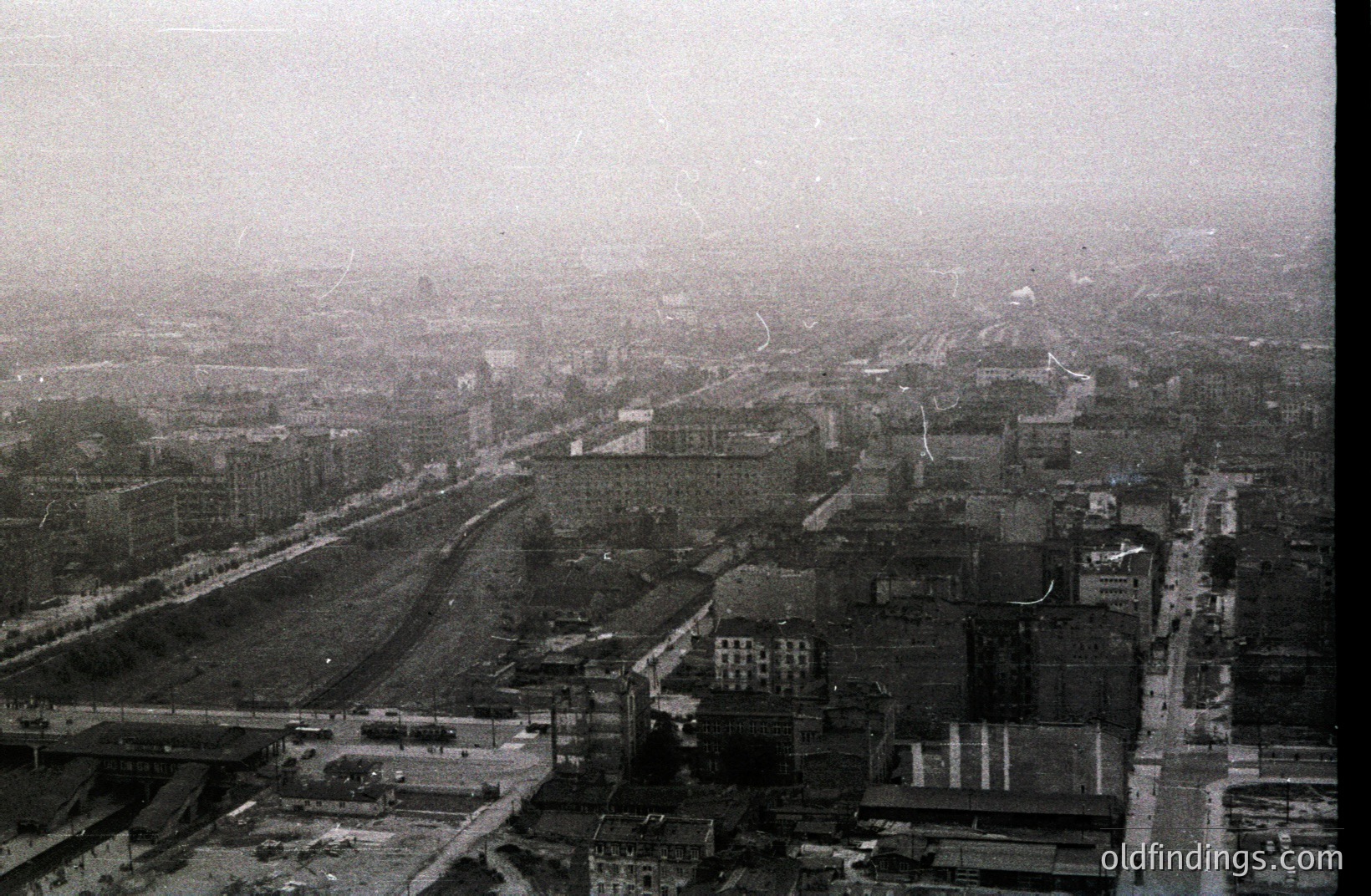 Overcast view of a city, likely industrial, with rail lines bisecting the scene. Dense atmospheric haze obscures distant structures and diminishes visibility. Appears to be a mid-20th century urban landscape, possibly post-industrial or undergoing change. Limited architectural details identifiable.