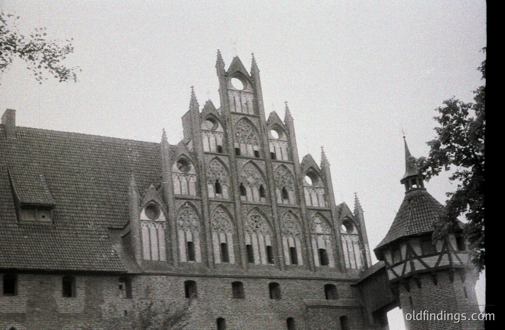 Striking Gothic-style brick facade of a medieval castle, showcasing intricate tracery and pointed arches. The roofline is steeply pitched with red tiles. A half-timbered tower is visible to the right. Likely a Northern European location.