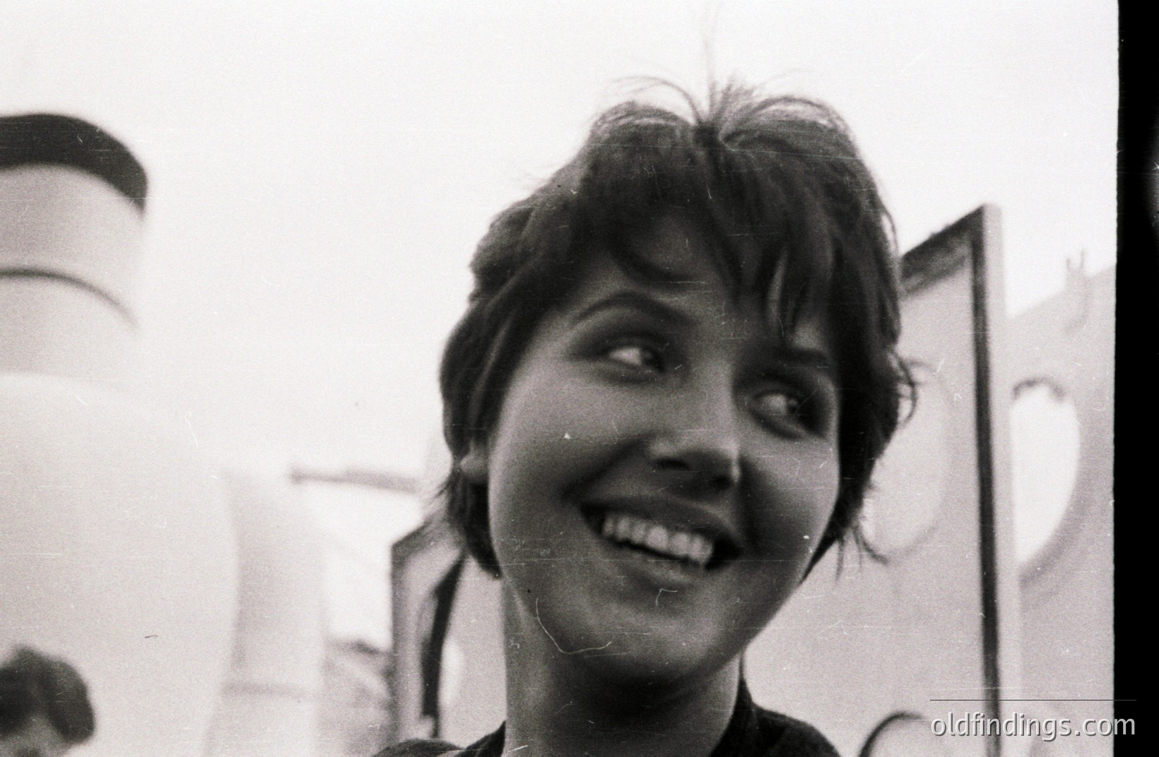Close-up portrait of a young woman with short, dark hair, smiling widely and looking off-camera. Grainy black and white image with visible film imperfections. Likely a candid snapshot from the 1960s or 70s, possibly taken outdoors given the background lighting. Simple composition, potentially a personal or family archive photograph.