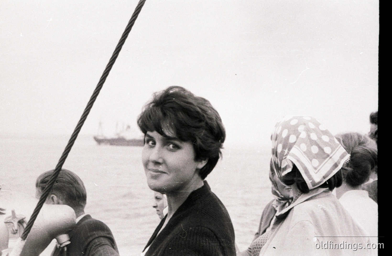 Black & white portrait of a young woman with short, dark hair, smiling toward the camera. Visible background features the ocean and a distant ship. Partial views of other individuals are included. Likely 1960s-1970s, coastal scene.