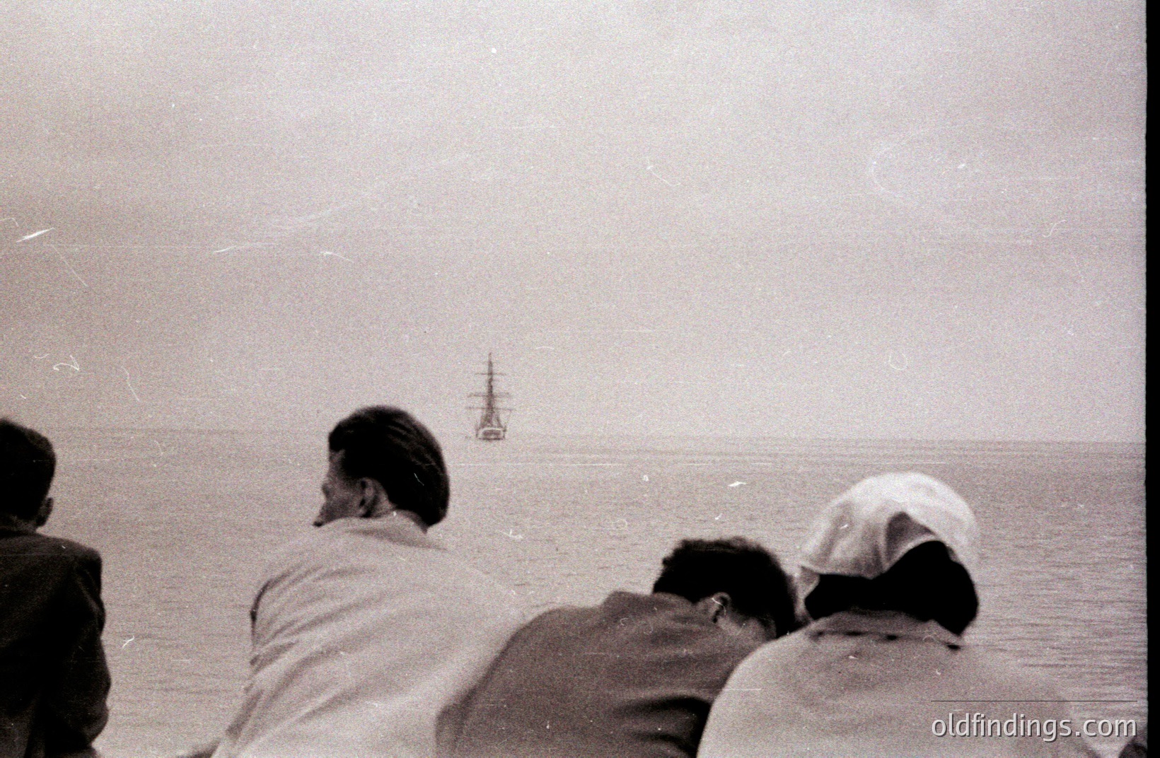 Three figures viewed from behind, possibly on a coastal overlook, gaze at a distant sailing ship on the horizon. The monochromatic image suggests an earlier era, possibly the mid-20th century. Grainy texture indicative of older film stock. A potentially poignant, contemplative scene.