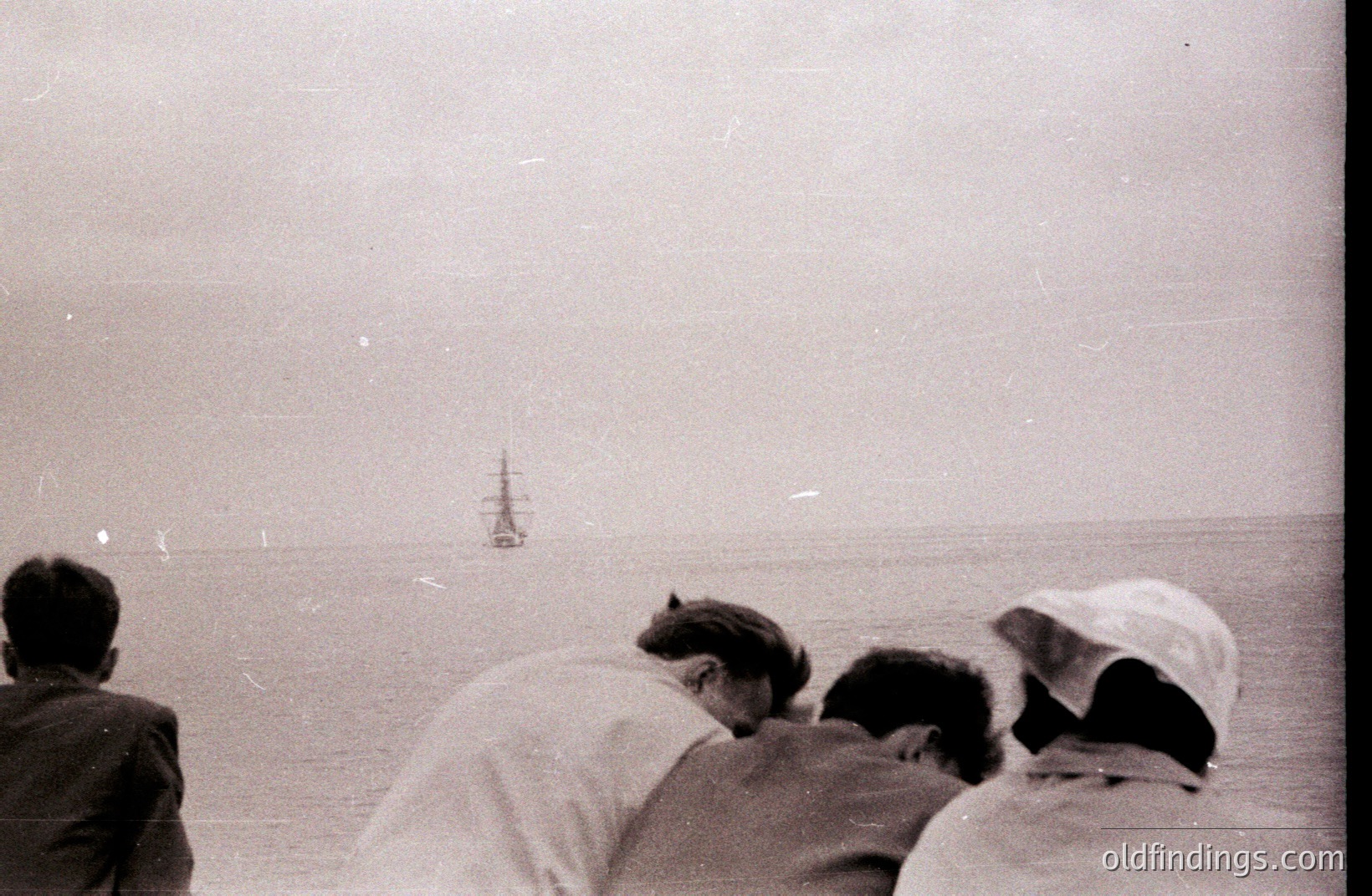 Three figures, likely children, are silhouetted against a hazy seaside view, intently observing a distant sailing ship. The monochromatic image suggests a nostalgic, possibly candid moment. Appears to be a coastal scene, potentially Eastern European. Grainy texture indicates older film stock.