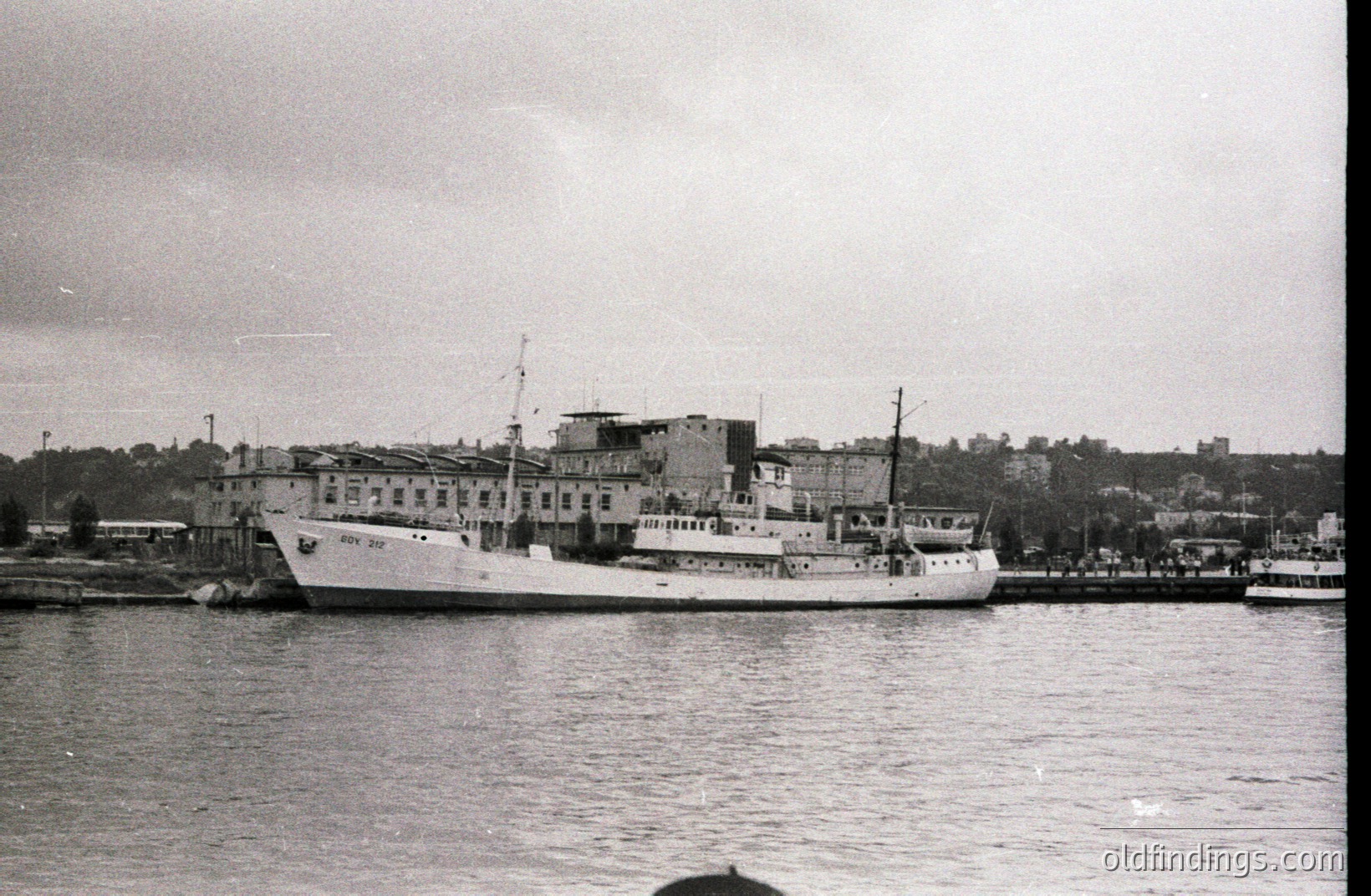 A coastal research vessel, "RV 312", is docked alongside a pier, viewed against a backdrop of a large, multi-story building, possibly a hotel or institutional complex. Likely taken in the 1960s or 70s, capturing a scene of maritime research and infrastructure. Visible harbor activity. Black and white photography.