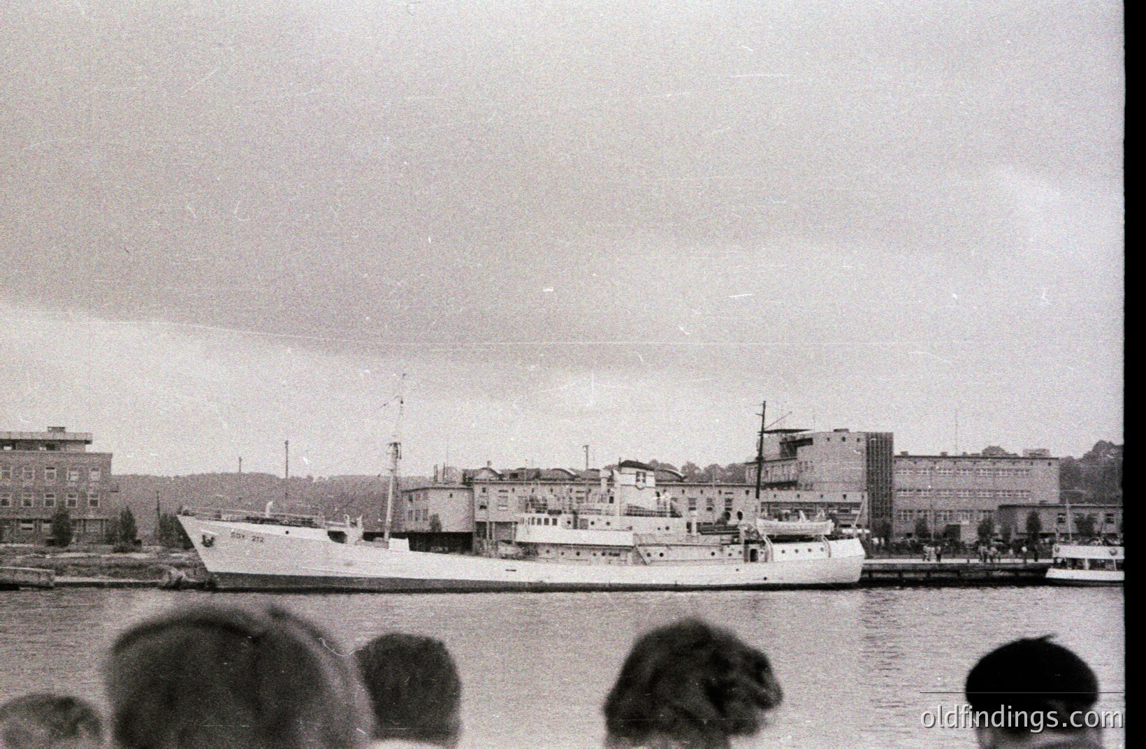 A medium-sized cargo ship is docked at a pier, framed by a multi-story industrial or administrative building. Partial view of spectators in the foreground. Appears to be a coastal town, possibly in Eastern Europe. Likely 1960s-1970s, based on photographic style & ship design.