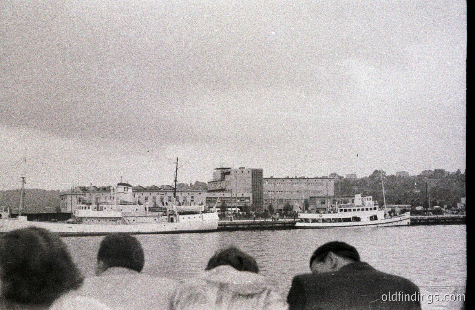 The harbor view depicts a coastal cityscape with two vessels at dock. Buildings feature modernist architecture and a sloping hillside backdrop. Likely Bulgaria, Varna; circa 1960s. The photo, taken from behind observers, captures a scene of everyday maritime activity. Potential value for historical research & design.
