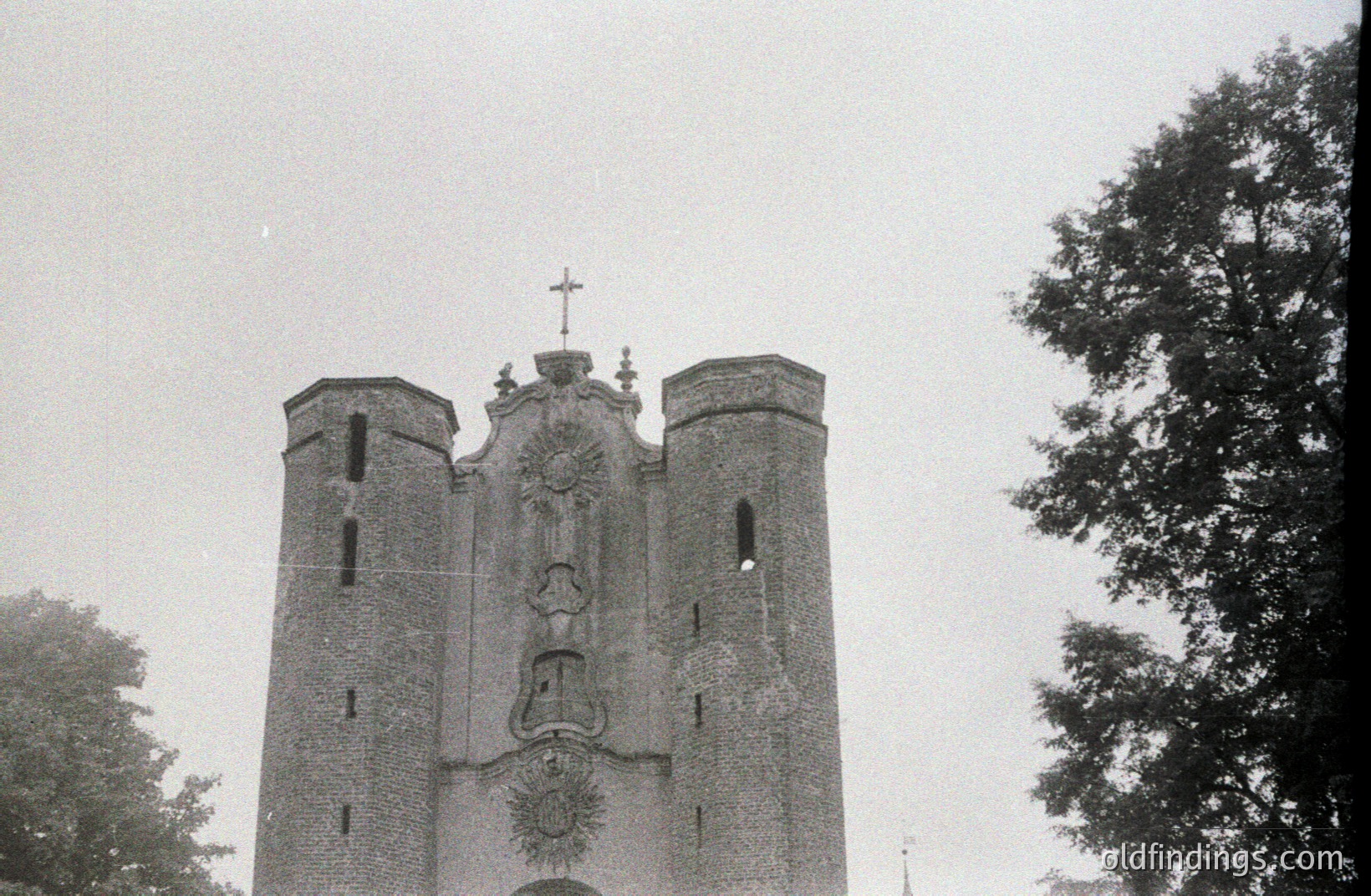 Striking architectural facade of a stone building, likely a church or civic structure. Features three central towers with a decorative crest above a recessed doorway. Tree branches frame the view. Appears to be a 1960s-1970s black and white photograph.