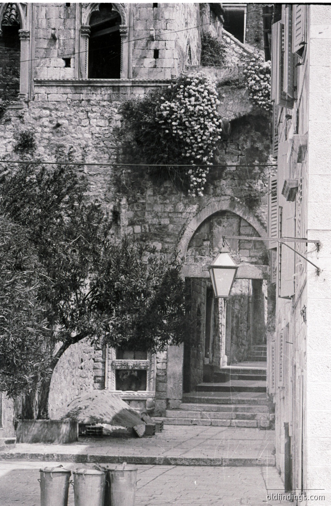 Stone steps lead up to an arched gateway framed by textured stone walls, overgrown with vegetation. A vintage street lamp illuminates the passage. Three metal barrels stand at the base of the stairs. Likely Southern Europe, early to mid 20th century.