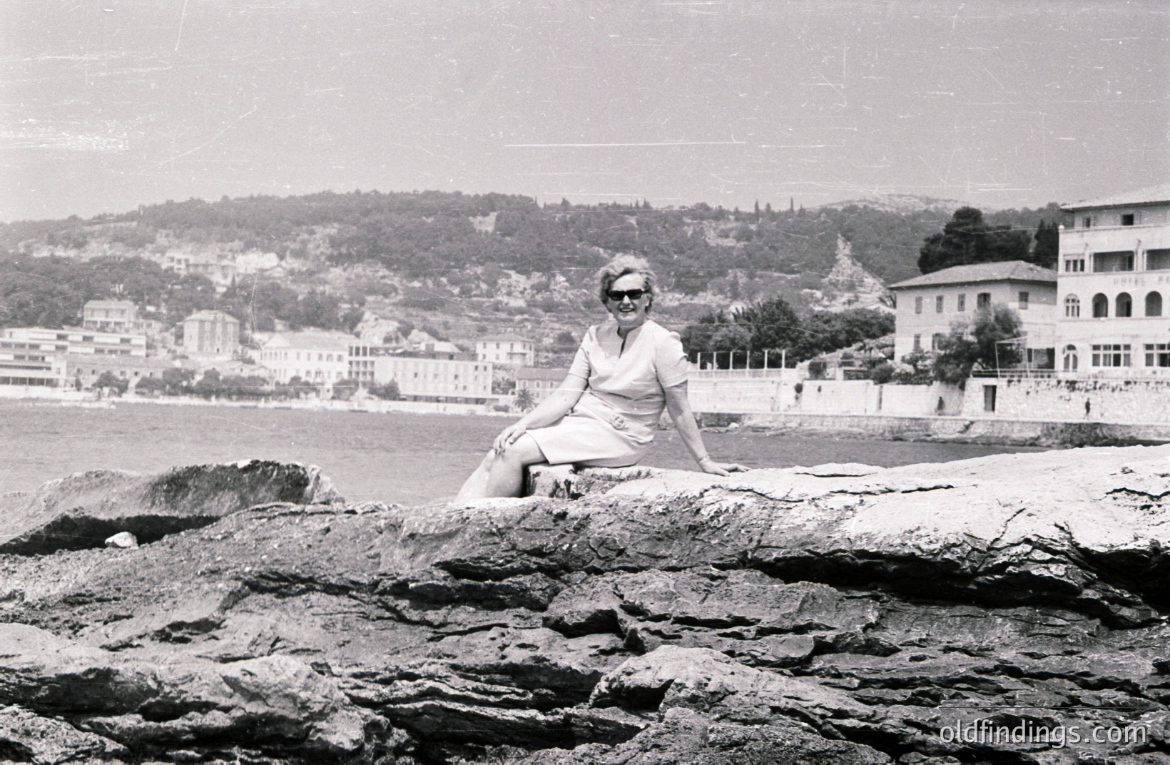 A woman in a knee-length dress and sunglasses sits casually on rocky coastline. Behind her, a seaside town with tiered buildings rises up a hillside, suggesting a Mediterranean location. Likely 1960s-1970s, captured in black and white. Charming, candid vacation moment.
