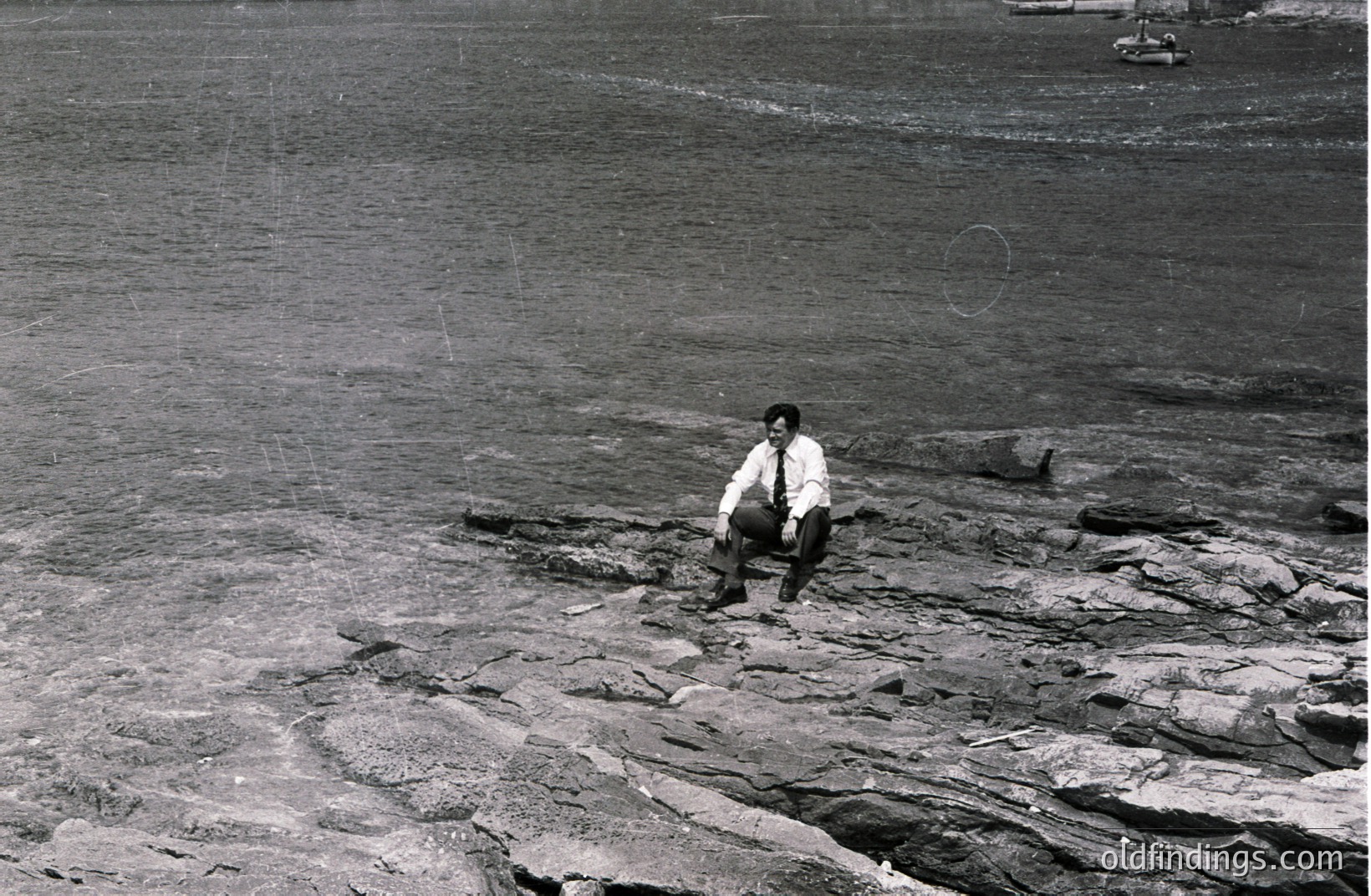 Man in a suit sits cross-legged on a rocky shoreline. The textured stone contrasts with dark water, likely a seaside or coastal location. His attire suggests a formal occasion or portrait from the mid-20th century, possibly the 1950s-1970s. Potential use for historical design or vintage aesthetic projects.